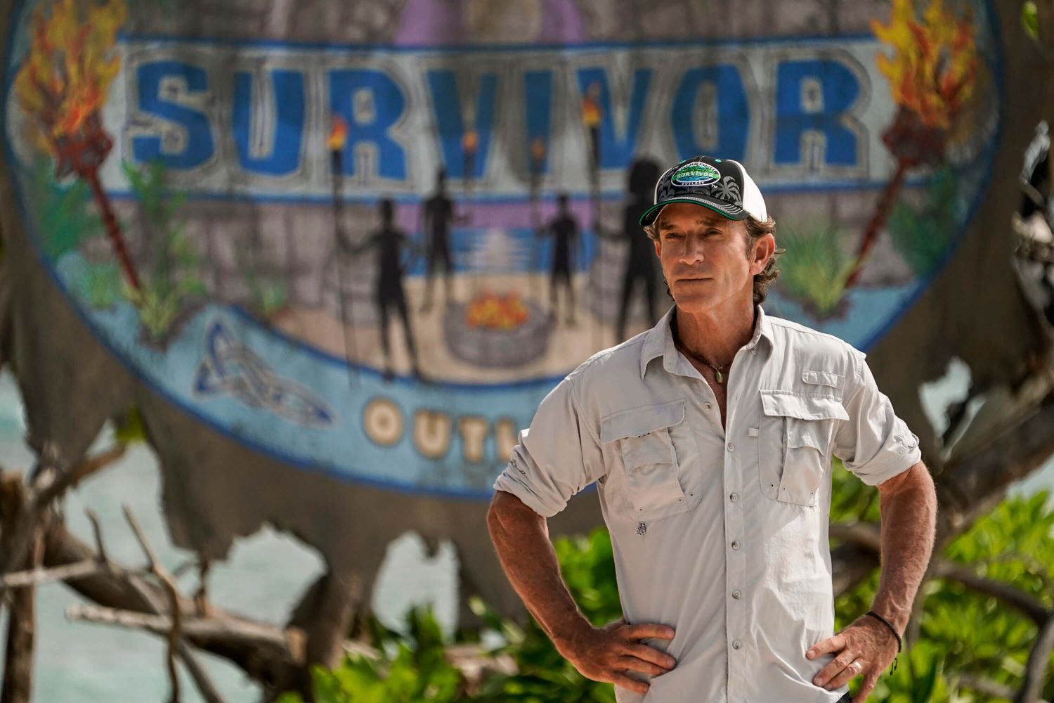 Jeff Probst, who previously confirmed the 'Survivor 44' showmance spoilers, wears a light gray button-up shirt with rolled-up sleeves and a black, white, and green 'Survivor' baseball cap.