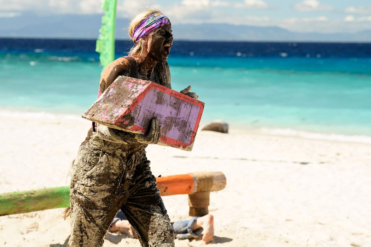 Carolyn Wiger competes during a Reward Challenge in 'Survivor 44' on CBS. Carolyn wears pants and a tank top caked in mud and sand while carrying a purple puzzle piece.