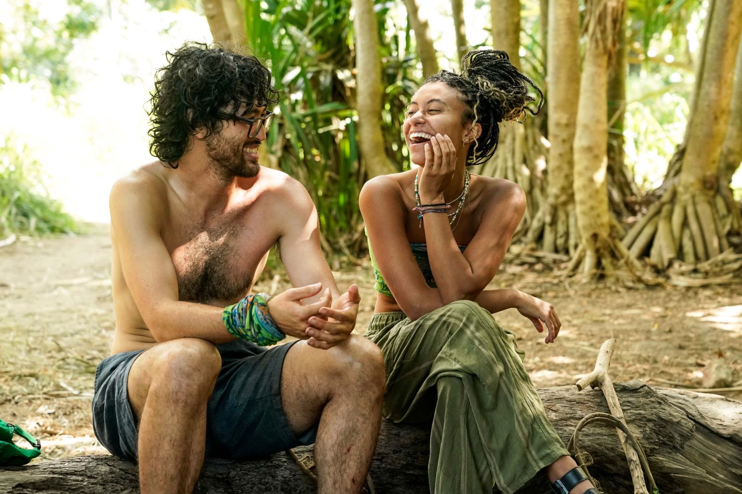 Matt Blankinship and Claire Rafson sit on a log at the Soka camp in 'Survivor 44' Episode 3, 'Sneaky Little Snake.' Matt wears dark gray shorts and his green 'Survivor' buff around his wrist. Claire wears her green 'Survivor' buff as a shirt and green pants.