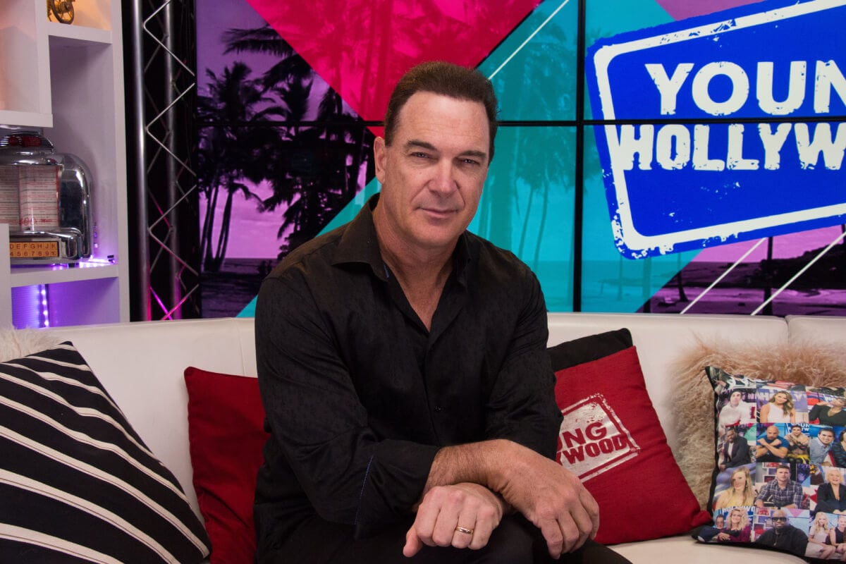 The Bachelor's uncle Patrick Warburton visits the Young Hollywood Studio. He sits on a couch wearing a black button-down shirt.