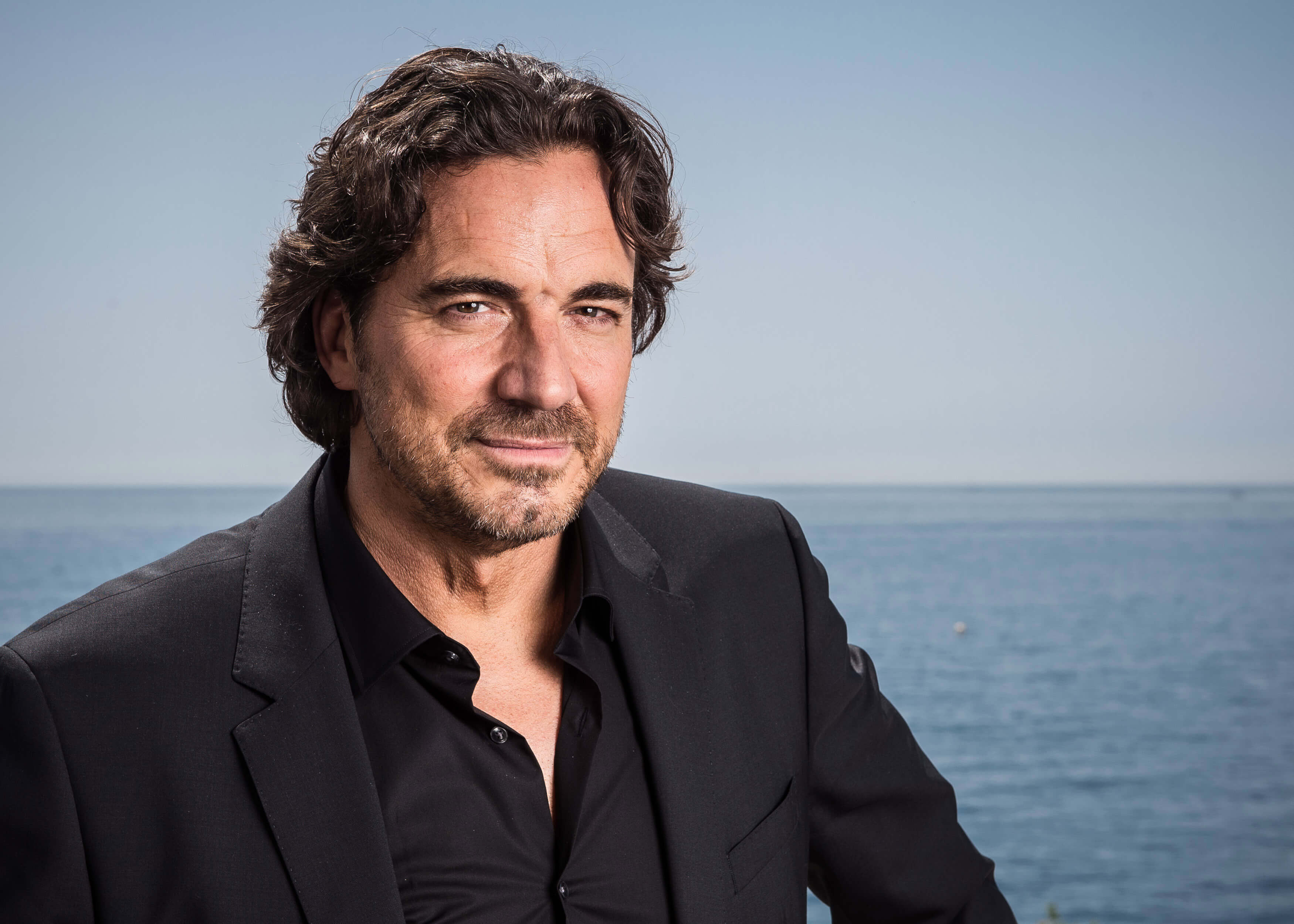 'The Bold and the Beautiful' star Thorsten Kaye dressed in a black suit; posing in front of an ocean backdrop.