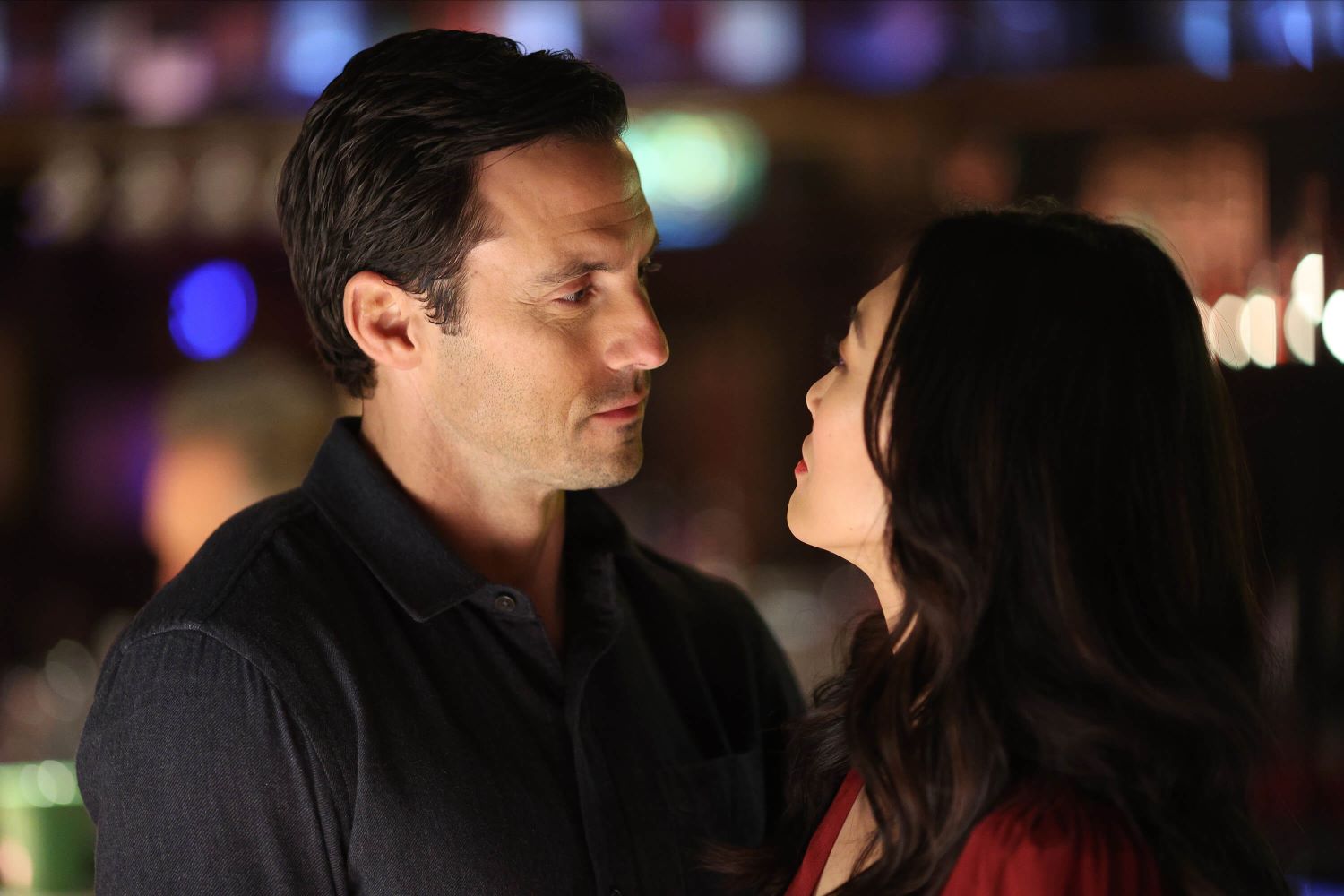 Milo Ventimiglia as Charlie Nicoletti and Catherine Haena Kim as Emma Hill in 'The Company You Keep' Season 1 Episode 3, 'Against All Odds.' Charlie wears a dark gray sweater, and Emma wears a red long-sleeved dress.