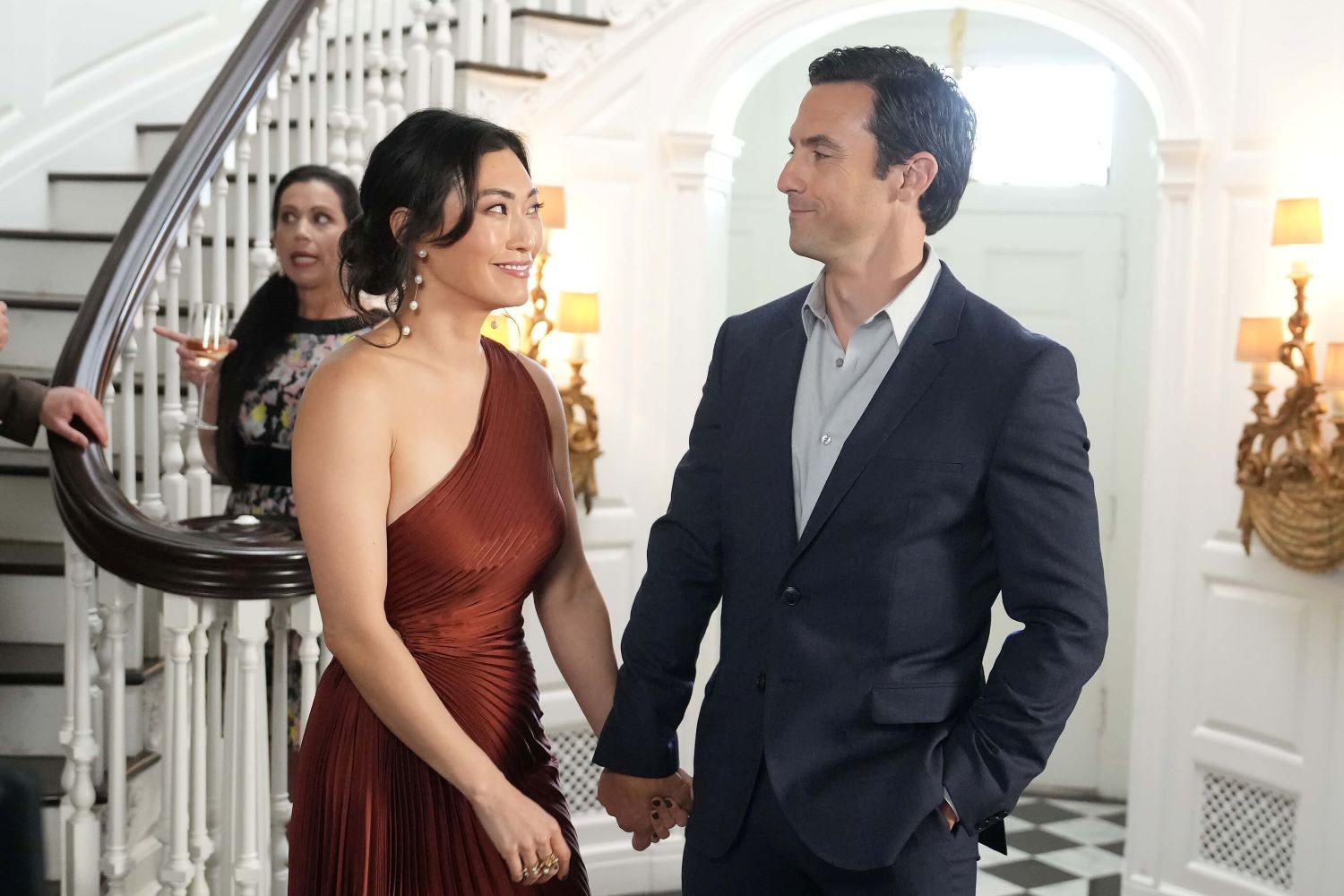 Catherine Haena Kim as Emma Hill and Milo Ventimiglia as Charlie Nicoletti in 'The Company You Keep' Season 1 Episode 4, 'All In.' Emma wears a reddish brown one-shoulder dress. Charlie wears a dark blue suit over a light gray button-up shirt.