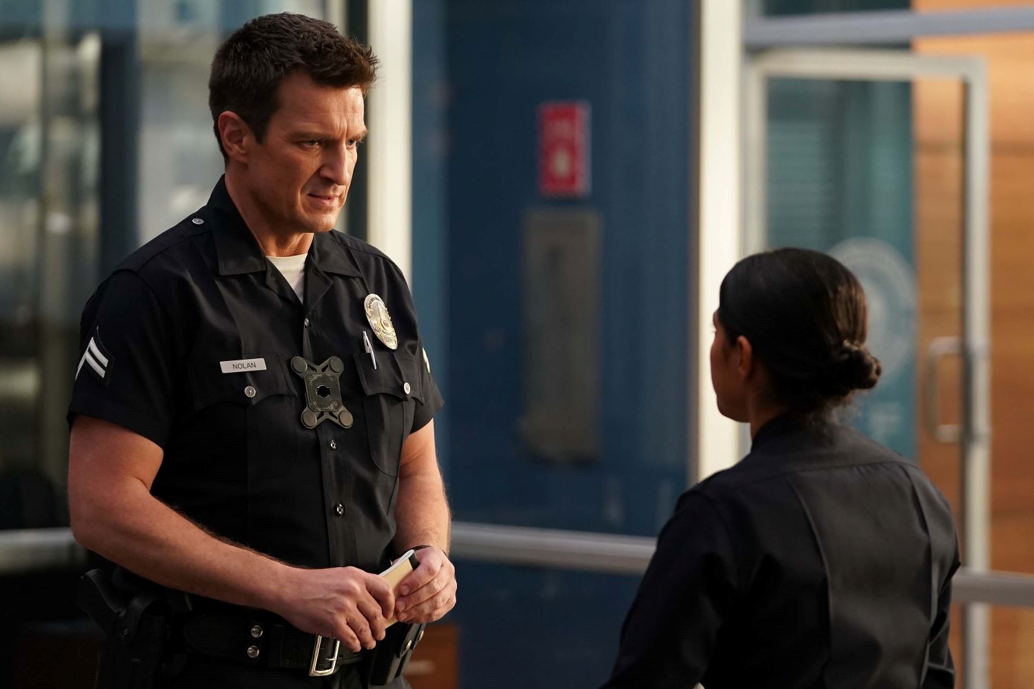 Nathan Fillion as John Nolan and Lisseth Chavez as Celina Juarez in 'The Rookie' Season 5 on ABC. Nolan and Celina talk to one another wearing their dark blue police uniforms.