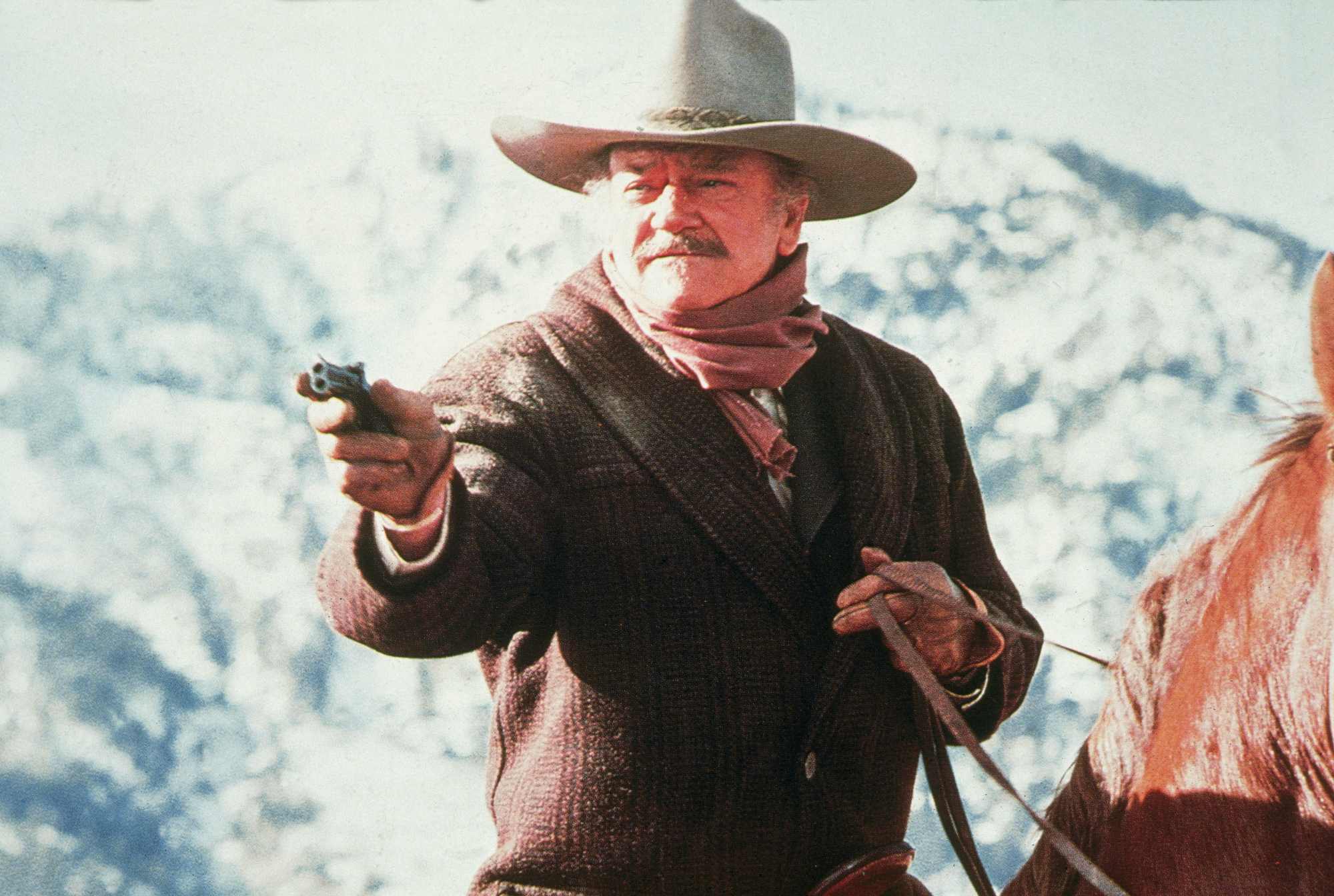 'The Shootist' John Wayne as J.B. Books holding out his pistol, while sitting on horseback.