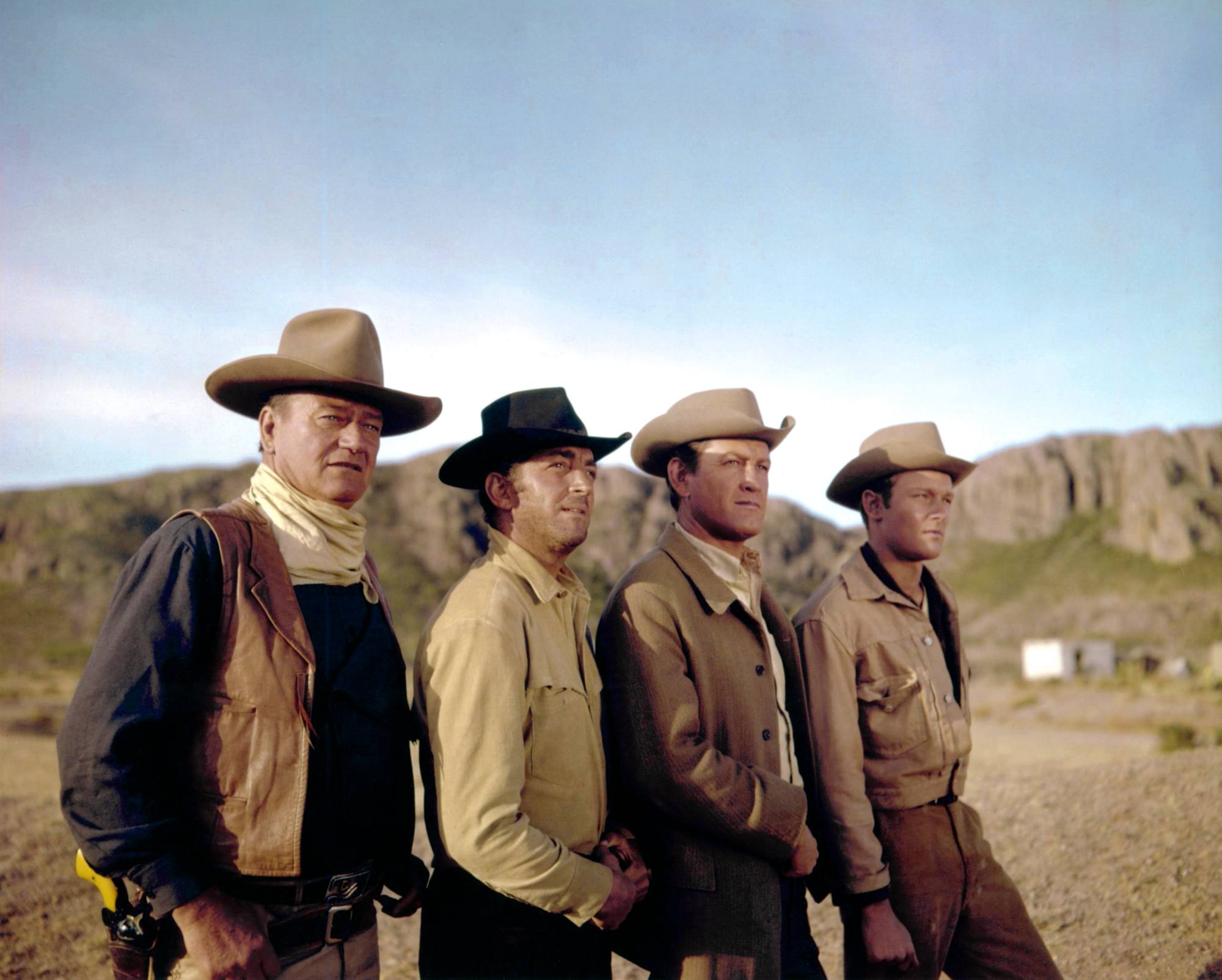 'The Sons of Katie Elder' John Wayne as John Elder, Dean Martin as Tom Elder, Earl Holliman as Matt Elder, and Michael Anderson Jr. as Bud Elder standing in a line wearing Western costumes in front of desert mountains.