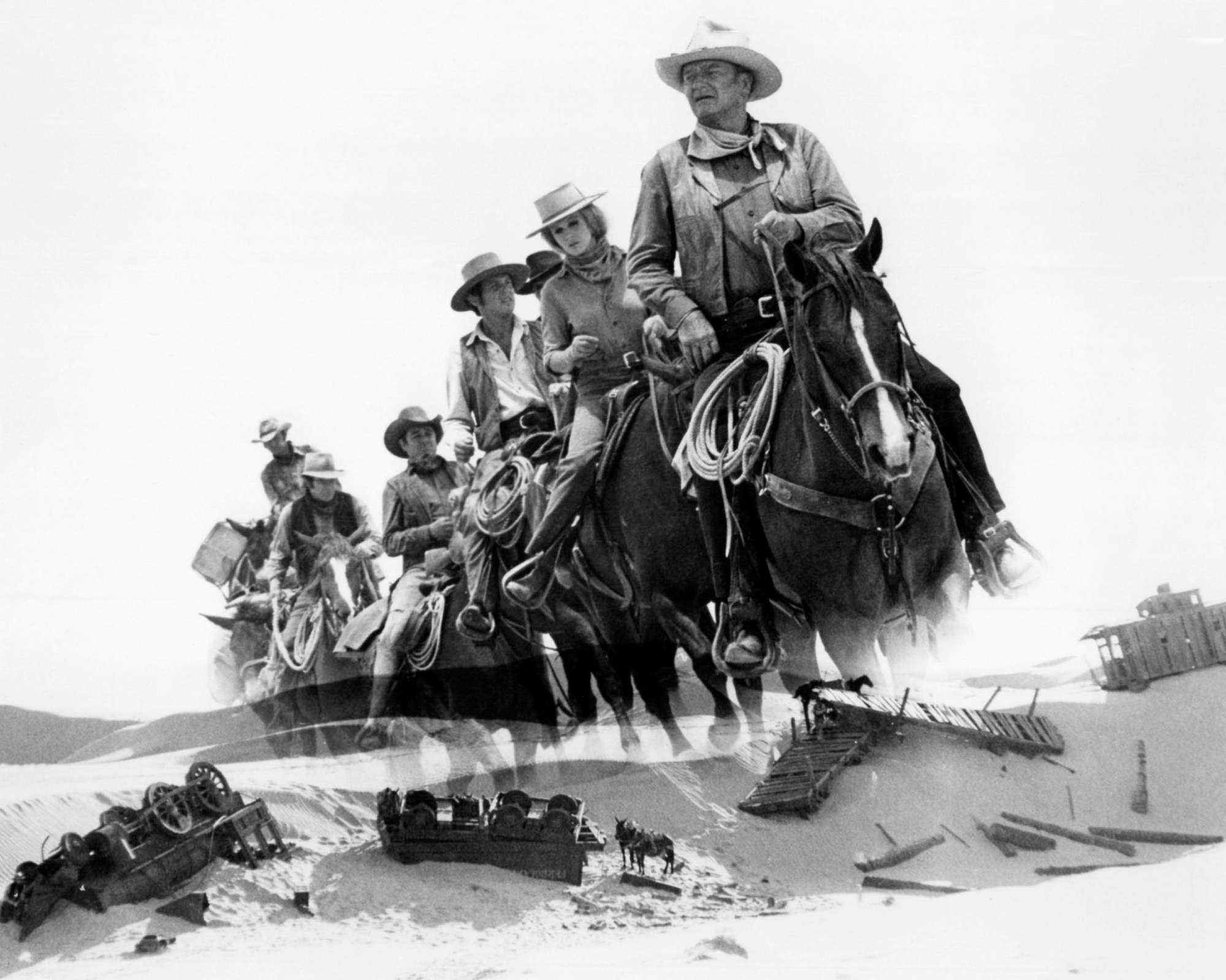 'The Train Robbers' John Wayne as Lane among other travelers on horses above an image of sand dunes with broken stagecoaches