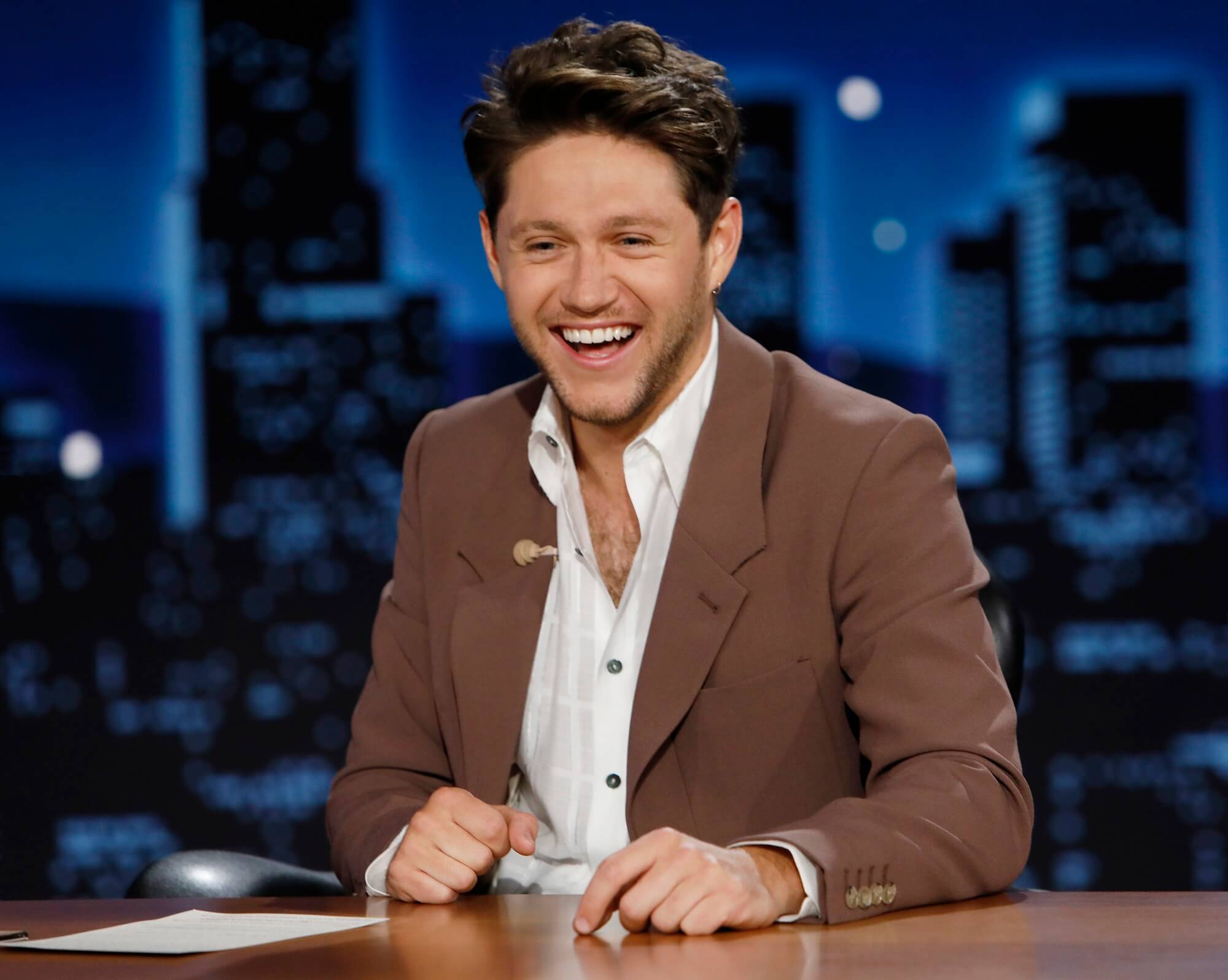 Niall Horan laughs while sitting at a desk in a white button down and brown jacket
