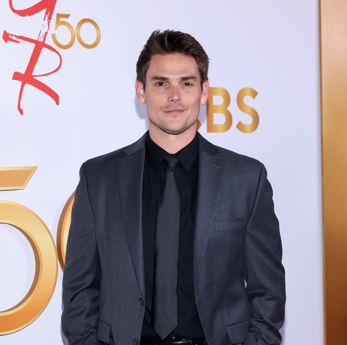 'The Young and the Restless' star Mark Grossman in a grey suit and black shirt; posing on the red carpet of the show's 50th anniversary celebration.
