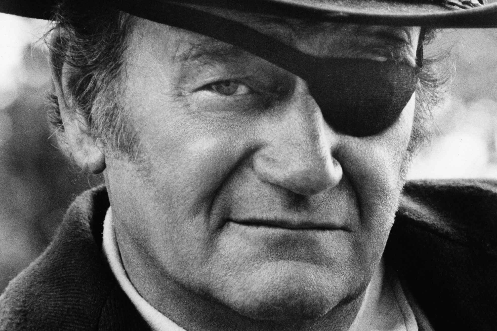 'True Grit' John Wayne as Rooster Cogburn wearing an eyepatch and cowboy hat with a slight smile on his face.