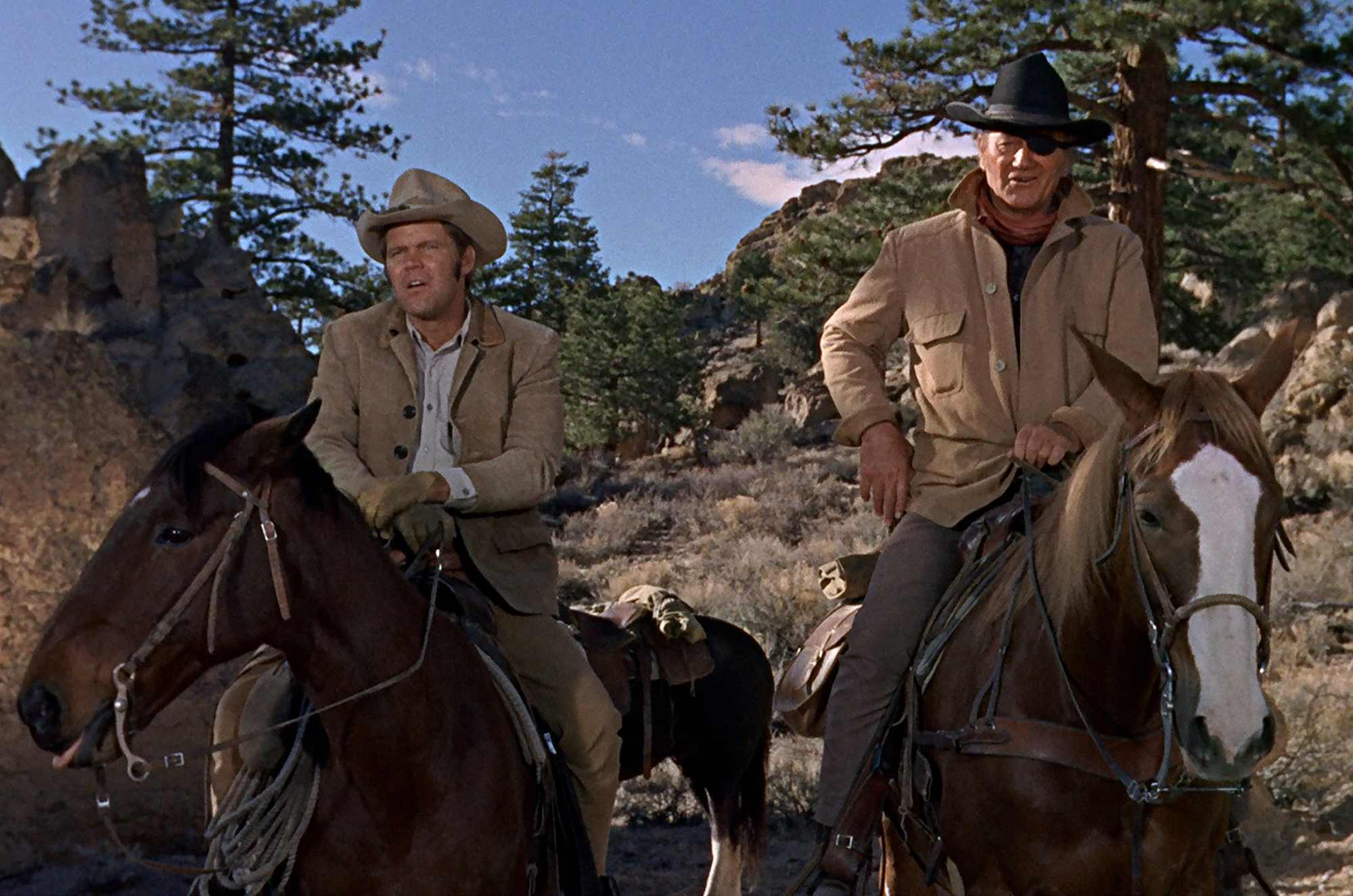 'True Grit' John Wayne as Rooster Cogburn and Glen Campbell as La Boeuf, each on a horse, standing in front of a mountain. Wayne has his hand on his waist.