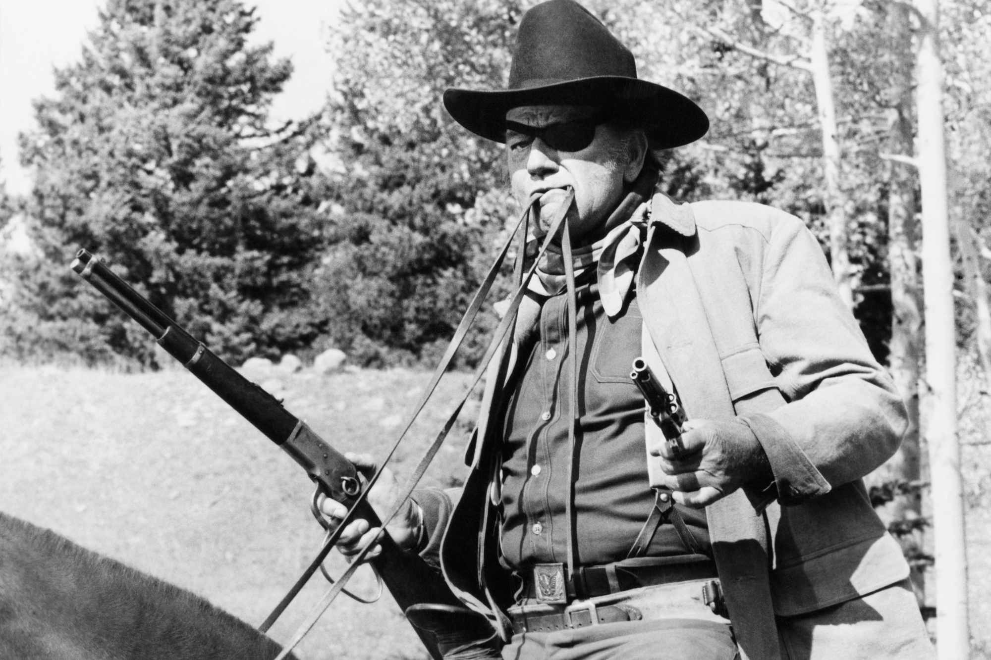'True Grit' John Wayne as Rooster Cogburn, with iconic quotes. He's on a horse, holding its reins in his mouth, while holding guns in his hands.