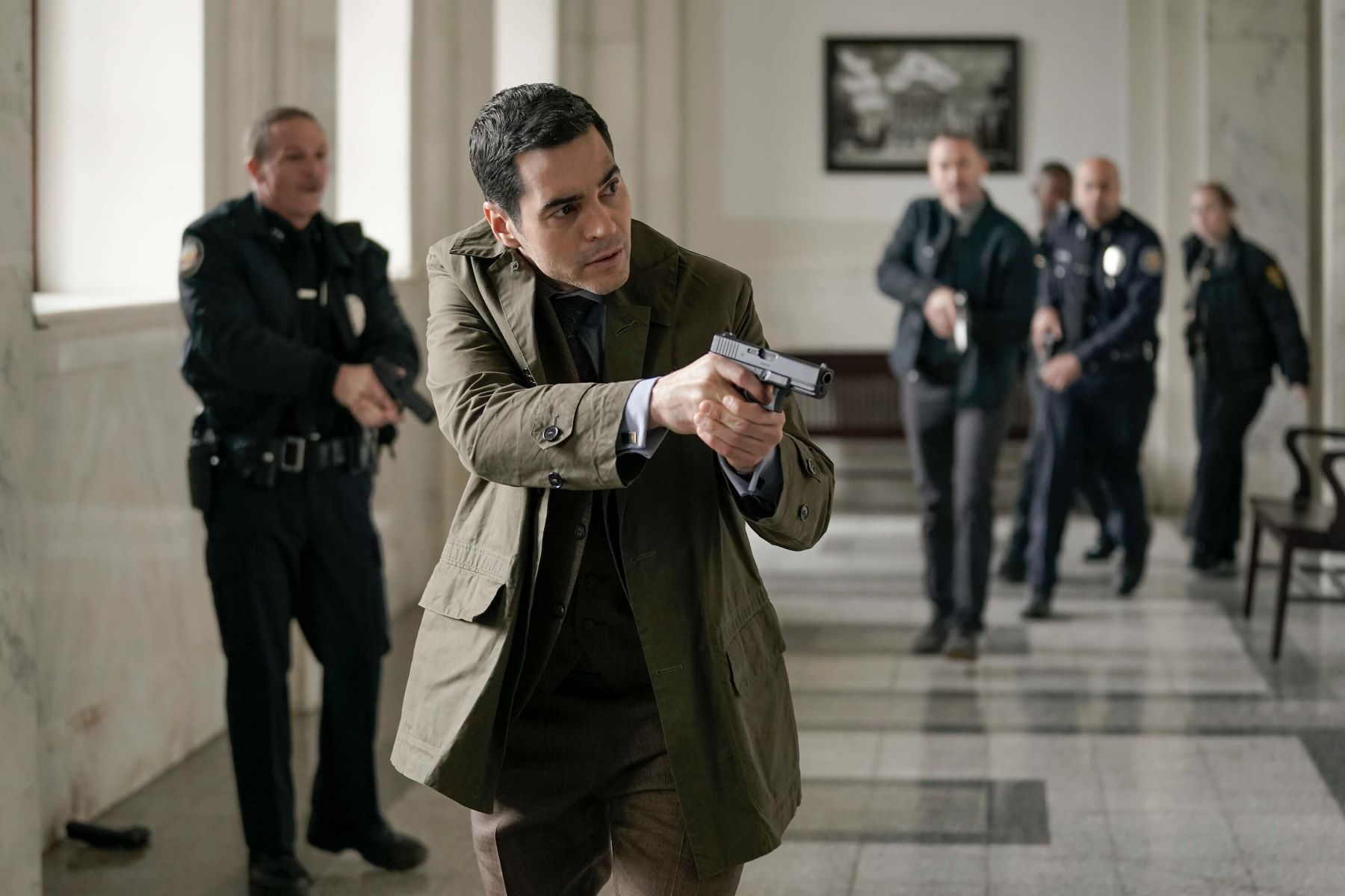 Ramón Rodríguez, in character as Will Trent in 'Will Trent' Season 1 Episode 9, 'Manhunt,' wears a dark green jacket over a gray three-piece suit while wielding a gun.