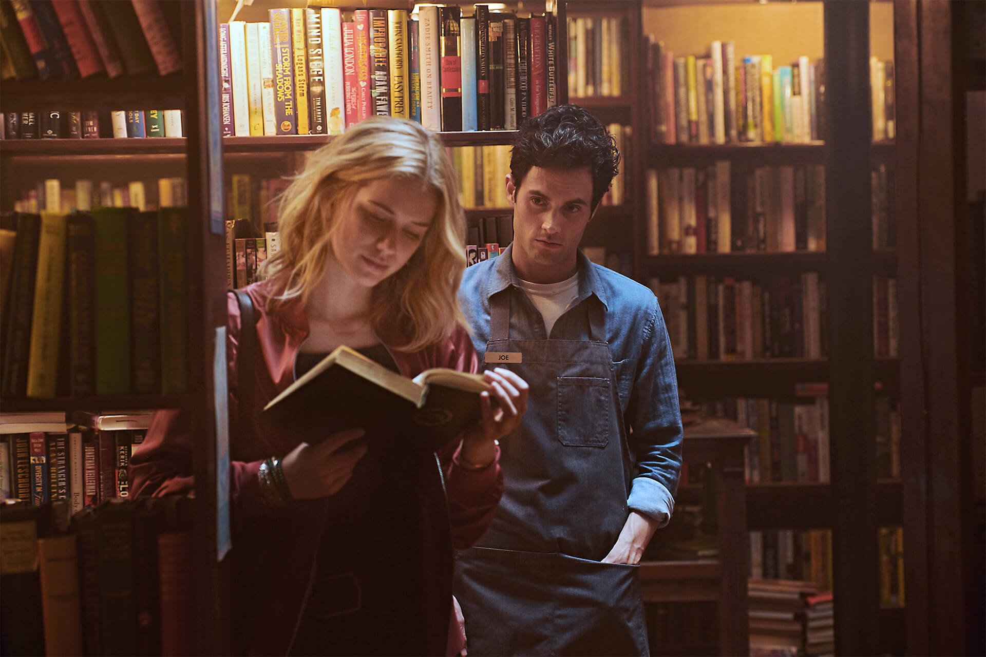 Penn Badgley as Joe Goldberg watches Elizabeth Lail as Guinevere Beck reading in the bookstore on 'You' Season 1.