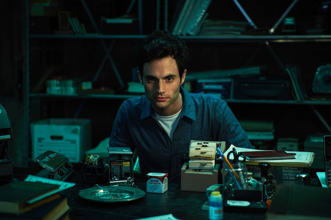 Penn Badgley as Joe Goldberg sits at a cluttered desk in the basement on 'You' Season 1.