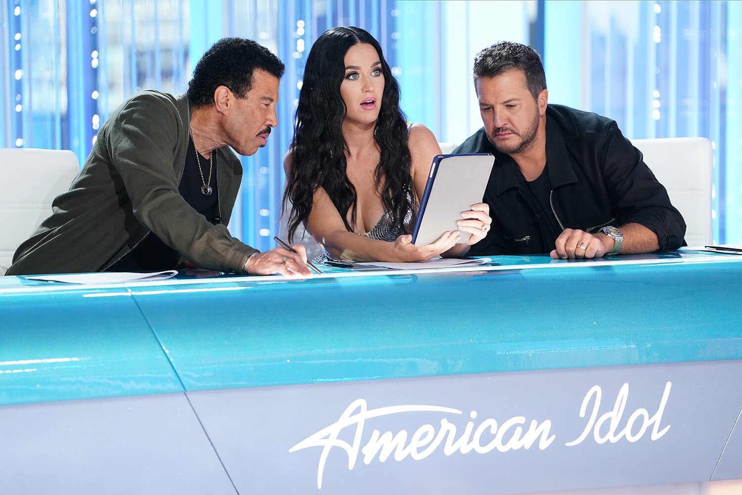 Lionel Richie, Katy Perry, and Luke Bryan lean over an iPad at the judges' table on American Idol