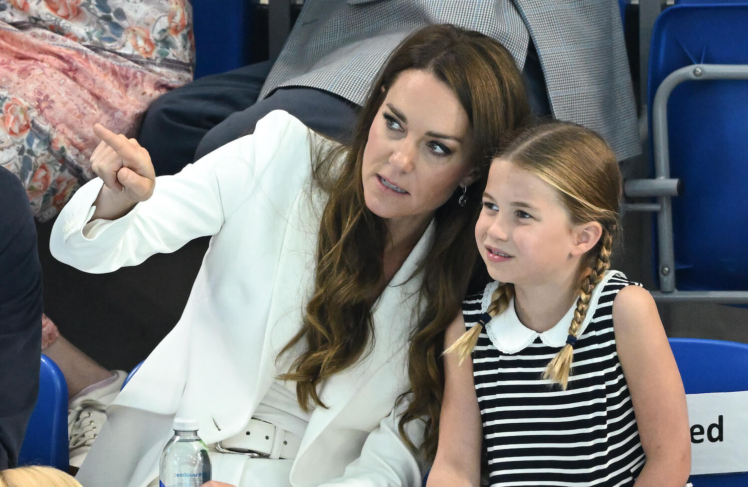 Kate Middleton wears a white jacket and points while talking to her daughter Princess Charlotte who smiles wearing black and white striped dress
