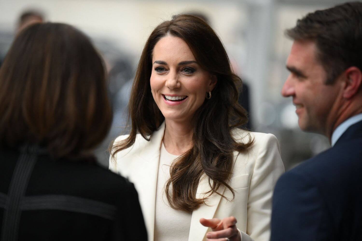 Kate Middleton smiles wearing a white suit jacket while talking with people
