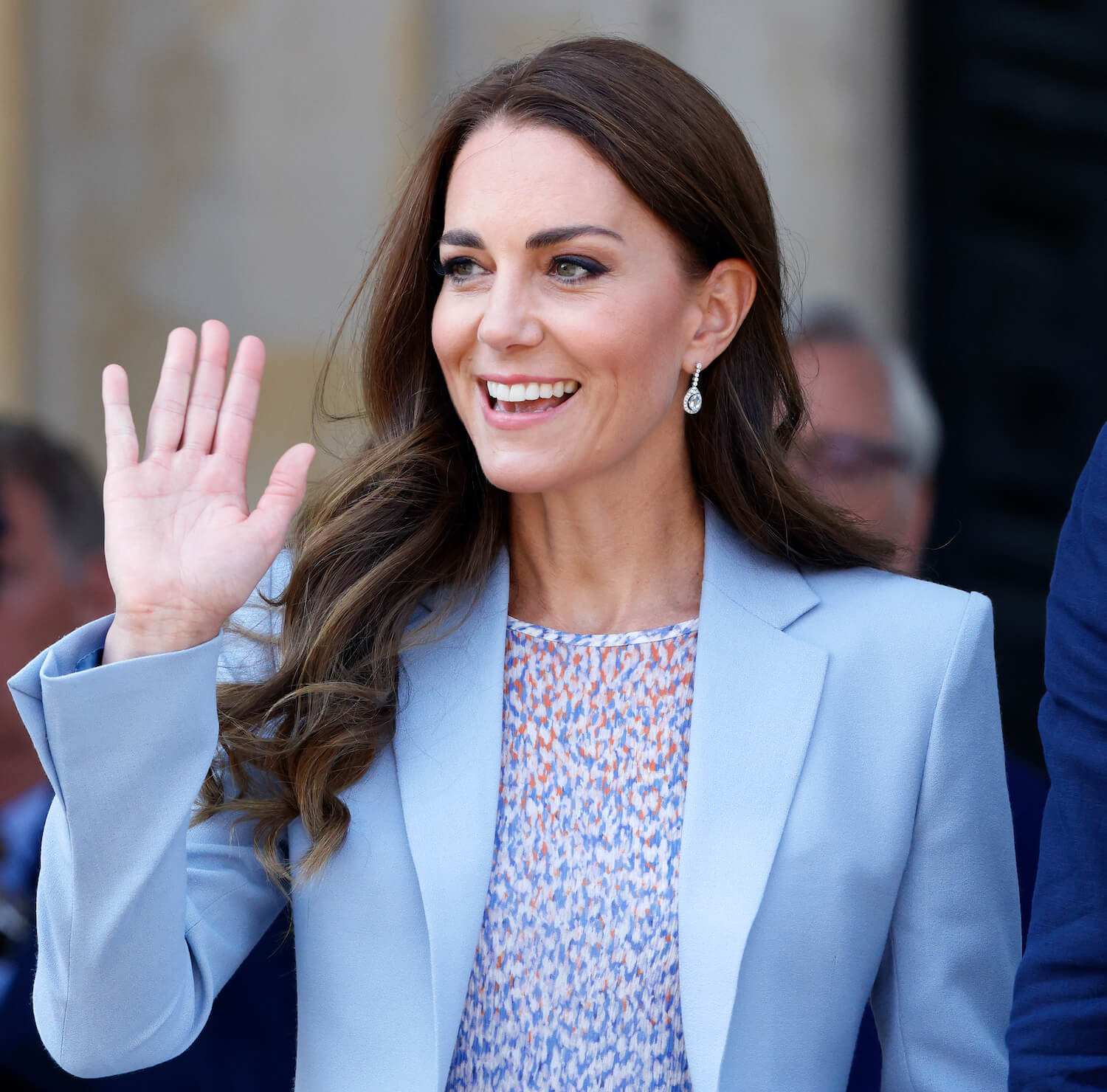 Kate Middleton sports natural hair and makeup and waves, wearing a light blue jacket
