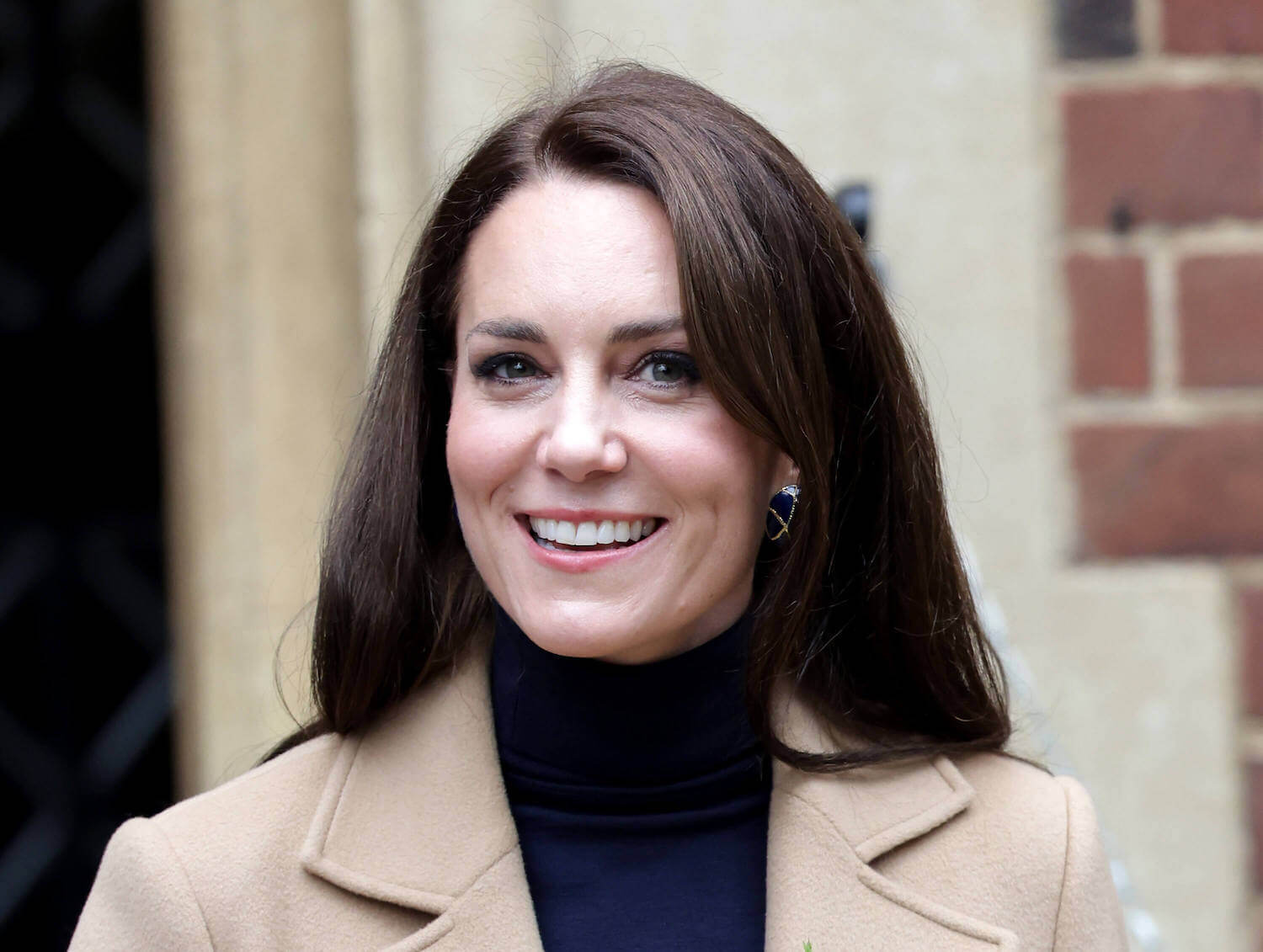 Kate Middleton wears natural makeup as she smiles wearing a black turtleneck and camel colored coat