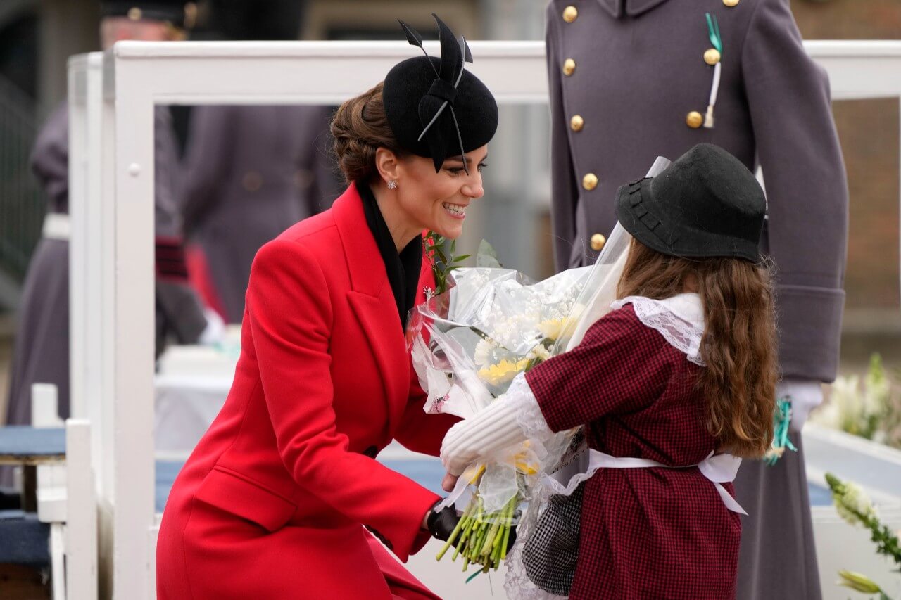 Kate Middleton, wearing a red Alexander McQueen coat, kneels down to greet a young well wisher.