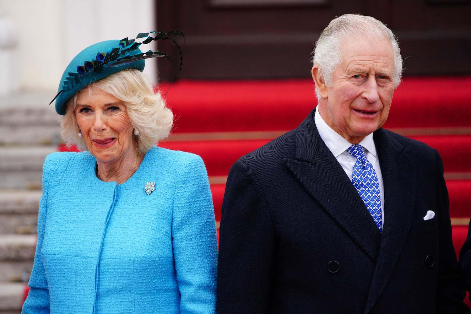 Camilla Parker Bowles wears a vibrant blue outfit with matching hat while standing next to King Charles dressed in a dark jacket and tie