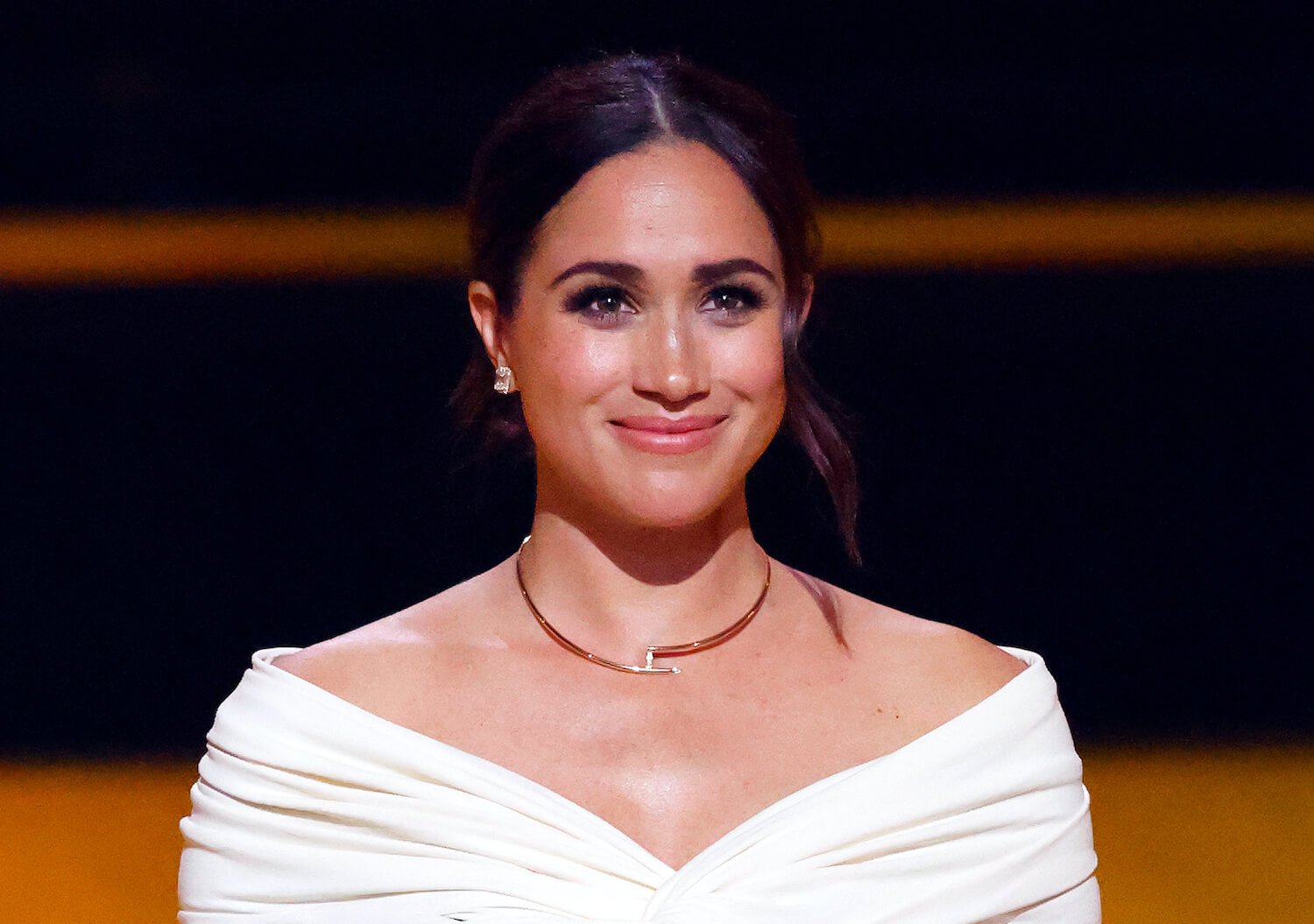 Meghan Markle smiles while wearing a white shoulder skimming top