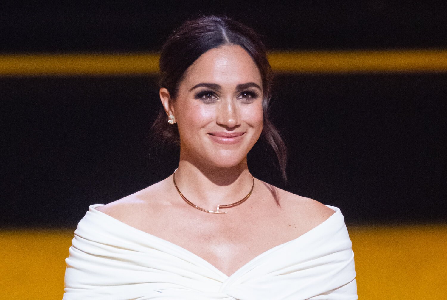Meghan Markle smiles while wering a white off the shoulder top