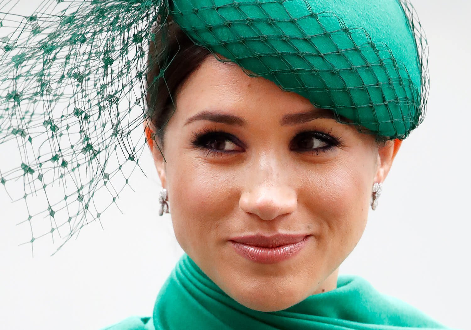 Meghan Markle gives a slight smile in a closeup shot of her wearing a green hat and matching green dress