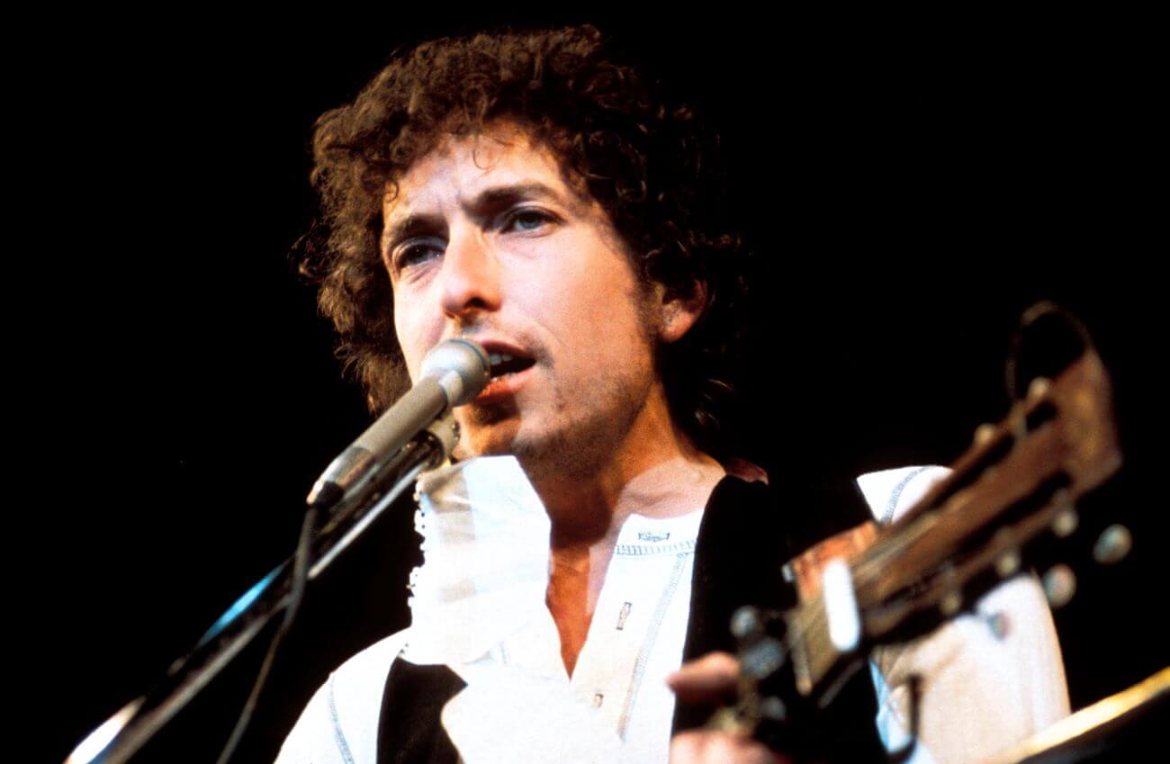 The musician Bob Dylan plays guitar and sings into a microphone.
