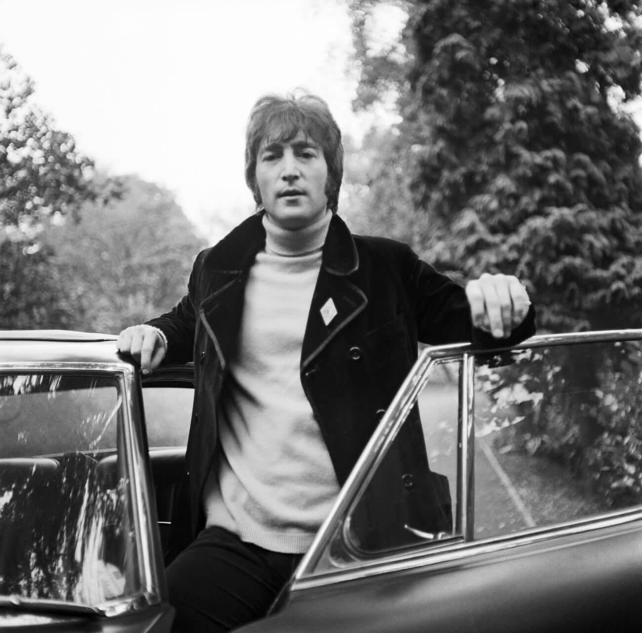 A black and white picture of John Lennon, one of the musicians in The Beatles, standing behind an open car door.