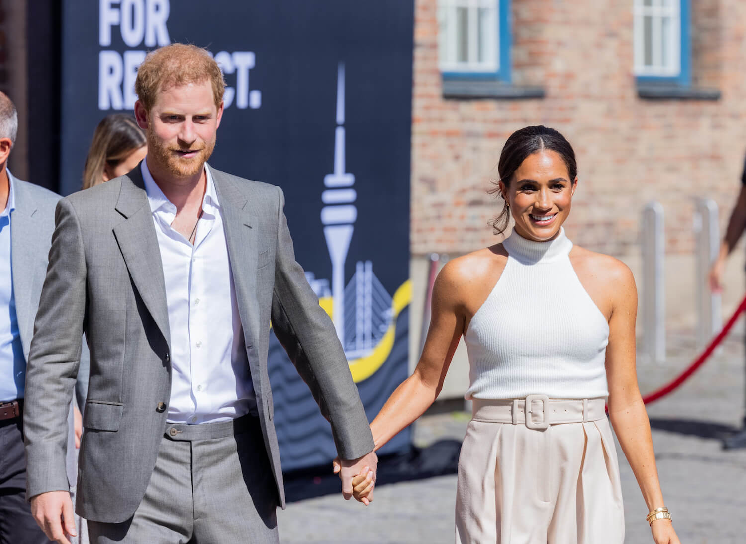 Prince Harry wears a gray suit and smiles while holding hands with Meghan Markle, dressed in tan pants and a white top
