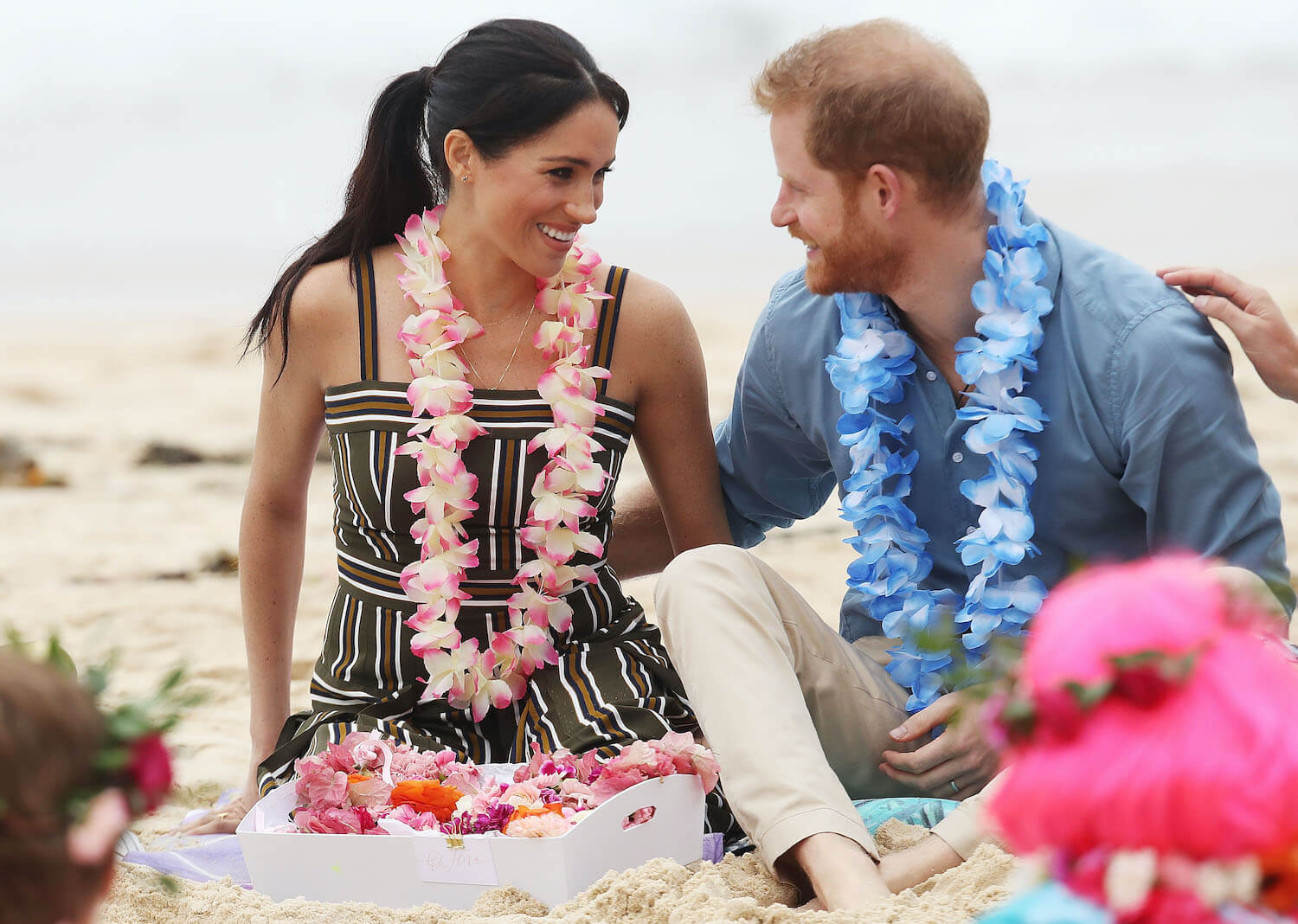 Prince Harry and Meghan Markle look at each other and smile while sitting on a beach wearing leis.