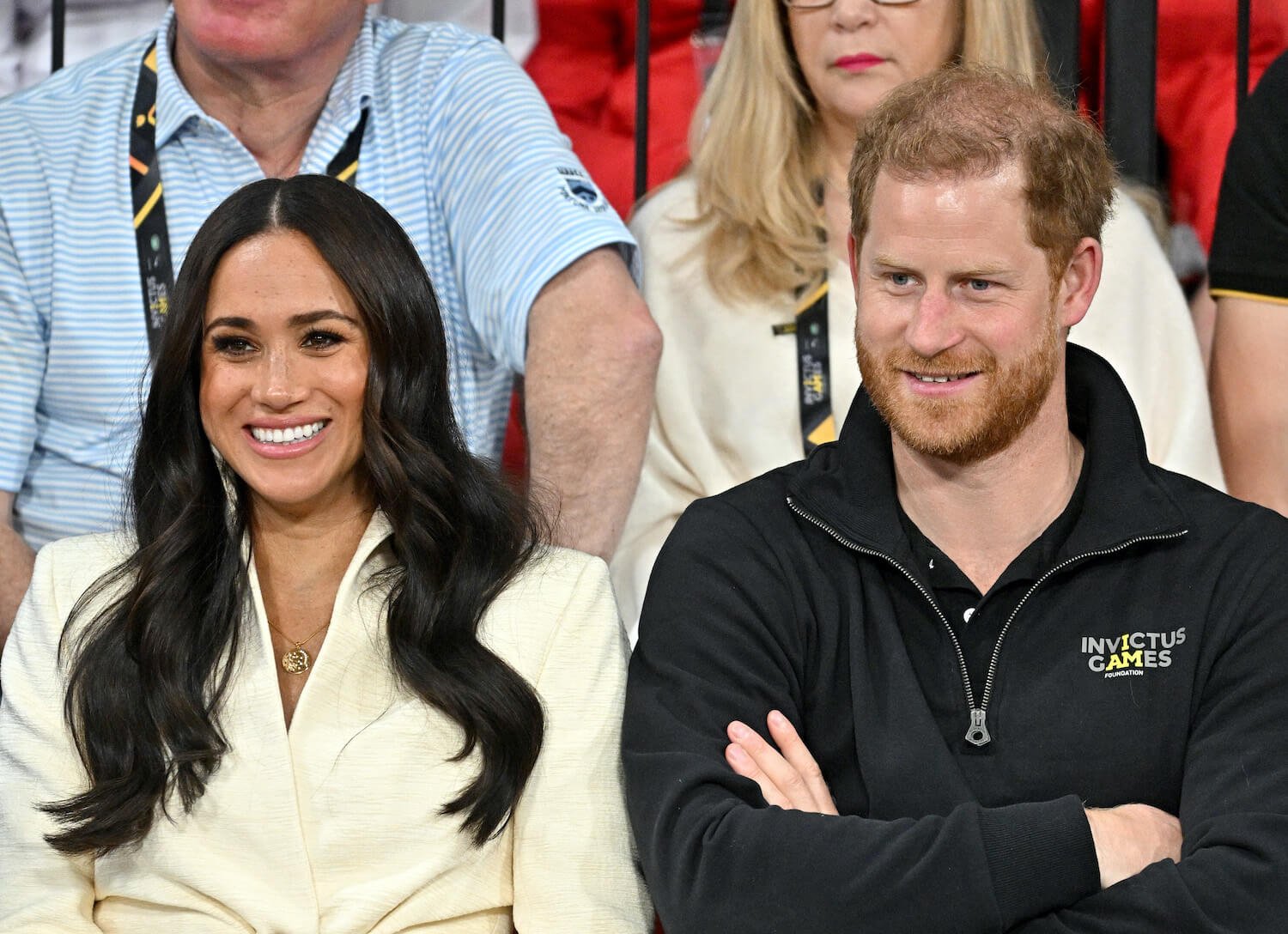 Meghan Markle wears whites and smiles while sitting next to smiling Prince Harry wearing a black top and crossing his arms