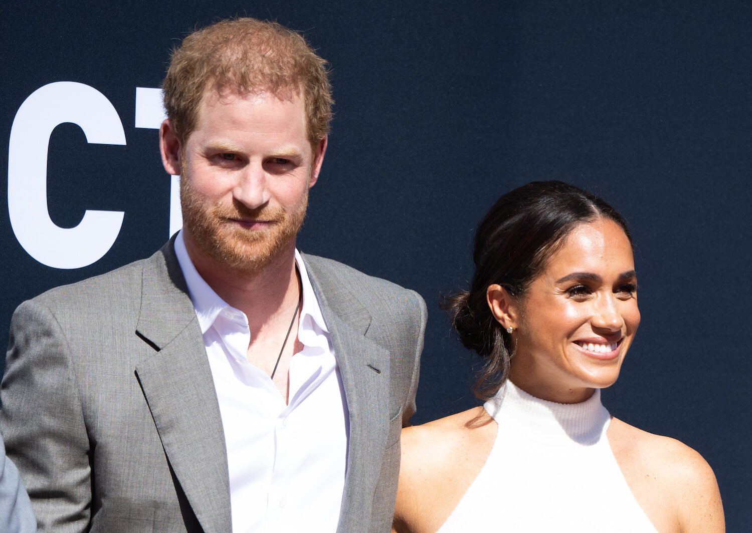 Prince Harry wears a jacket and white shirt and smirks while standing next to Meghan Markle, who is smiling while wearing a white top