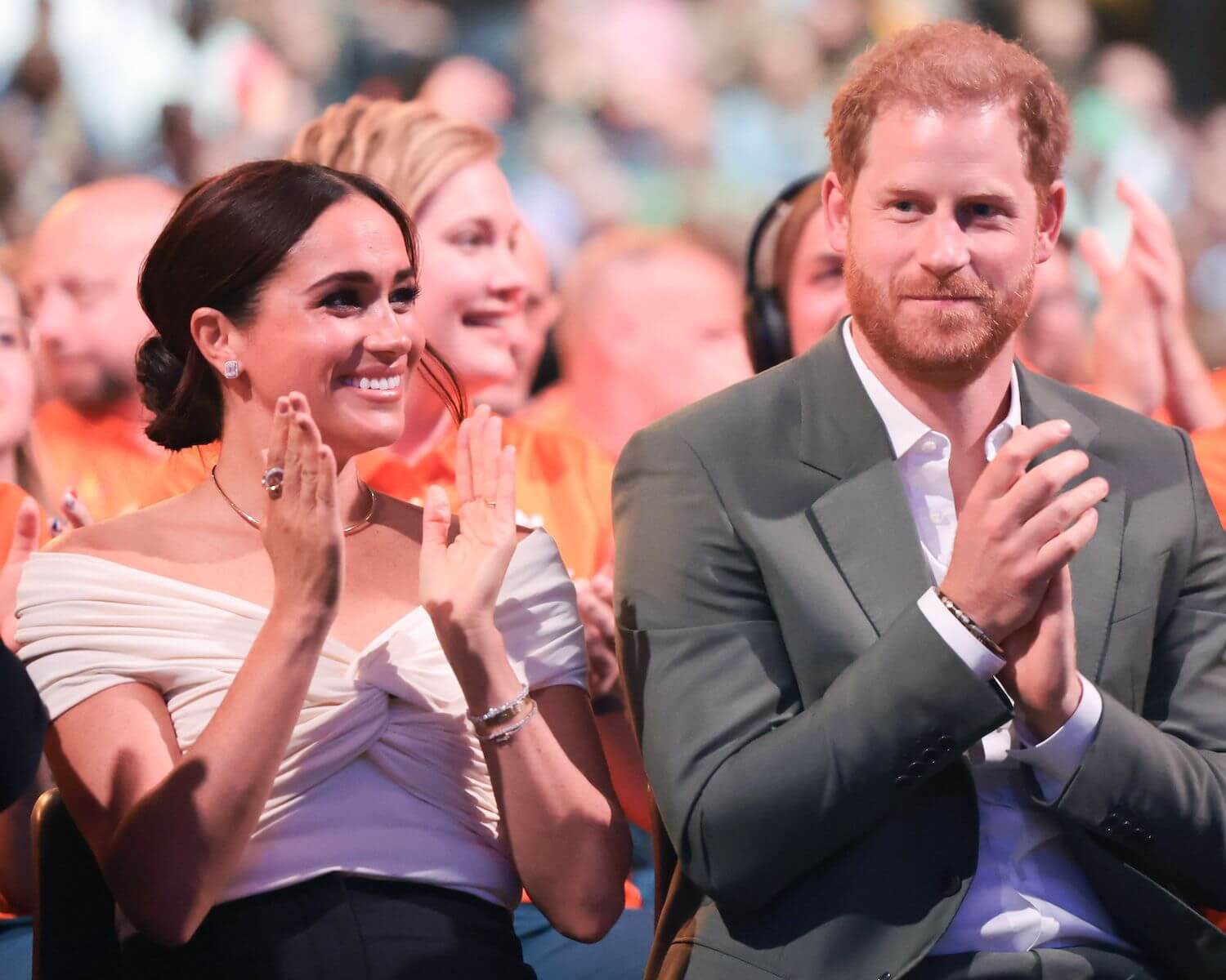 Meghan Markle wears a white off the shoulder top while sitting next to Prince Harry as both smile and clap their hands