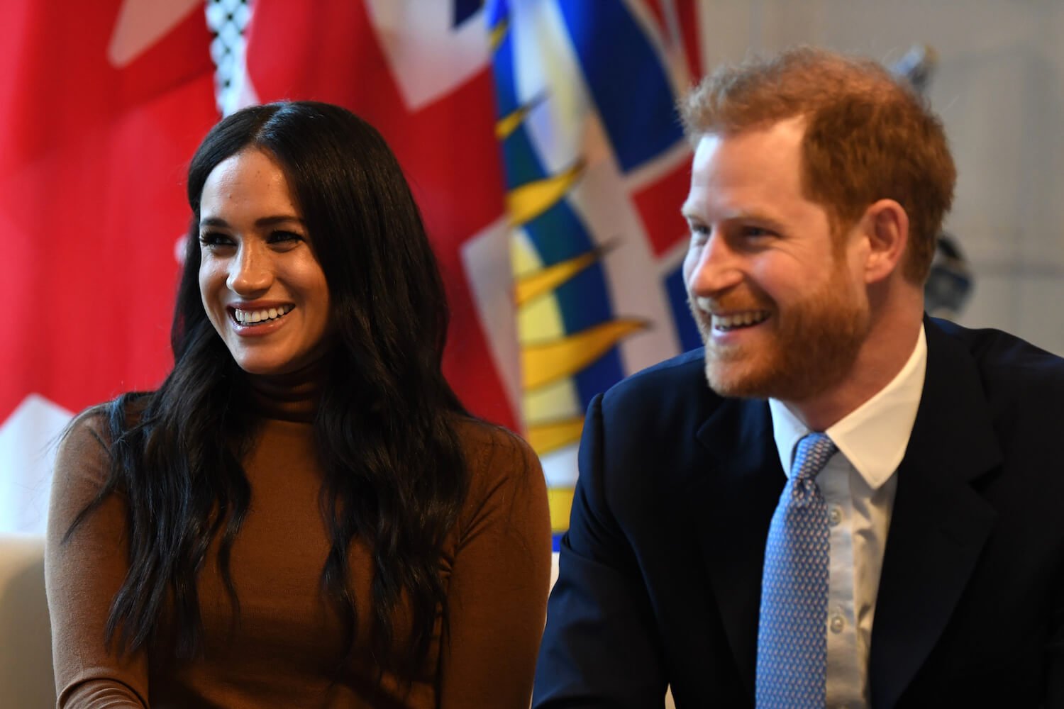 Meghan Markle wears a brown turtleneck and smiles while sitting next to Prince Harry, wearing a suit and tie and smiling