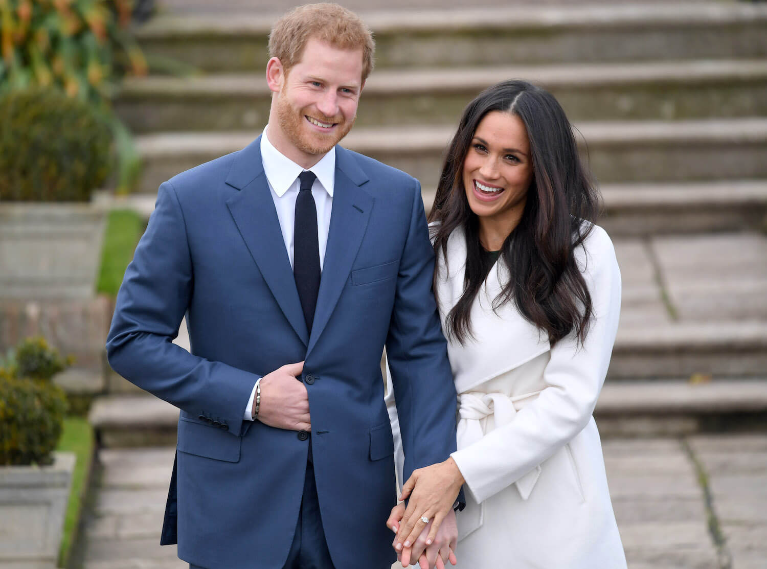 Prince Harry wears a suit, smiles, and holds hands with Meghan Markle who smiles while wearing white and showing off her engagement rings