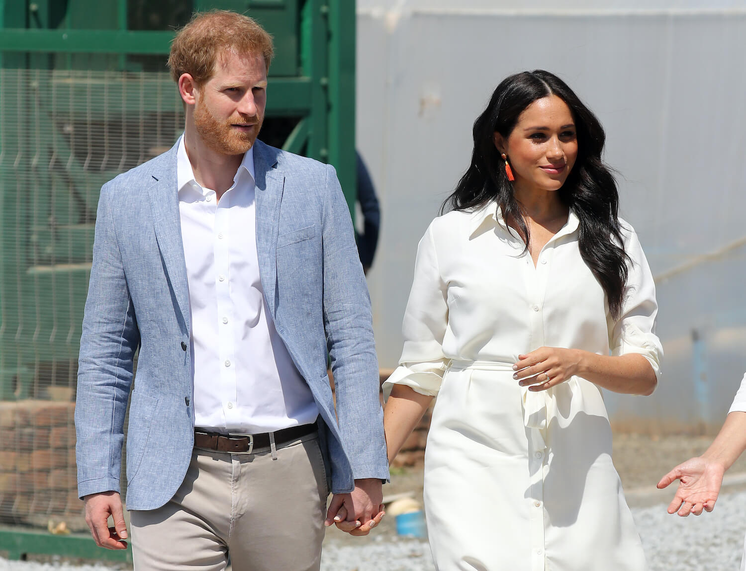 Prince Harry wears a light blue jacket and holds hands with Meghan Markle, wearing a white dress