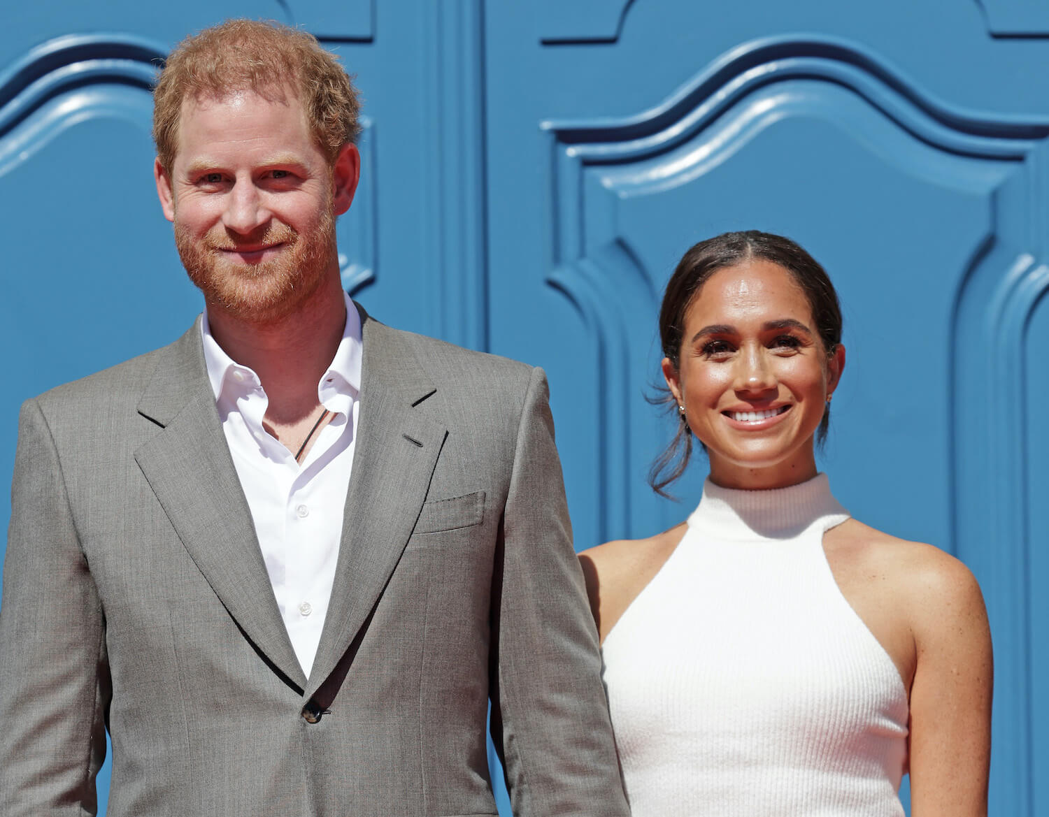 Prince Harry and Meghan Markle smile while standing in front of a blue door, Meghan wearing a white top and Harry wearing a white shirt and jacket