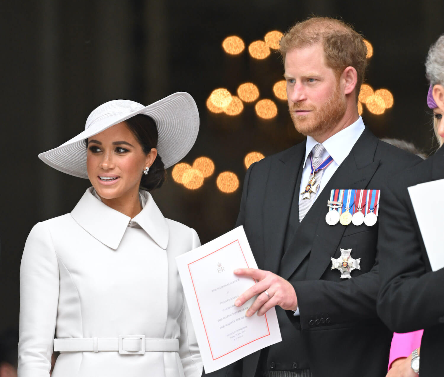 Meghan Markle wears a white coat and white hat, while Prince Harry wears a suit and tie as they both look on