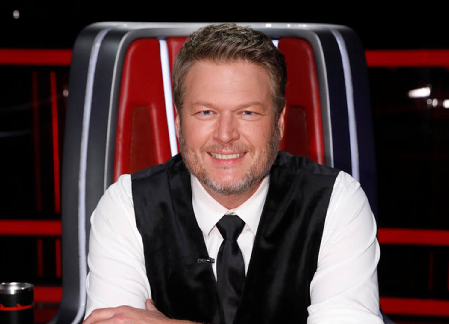 Blake Shelton sits in his chair and smiles while wearing a black vest and tie on The Voice