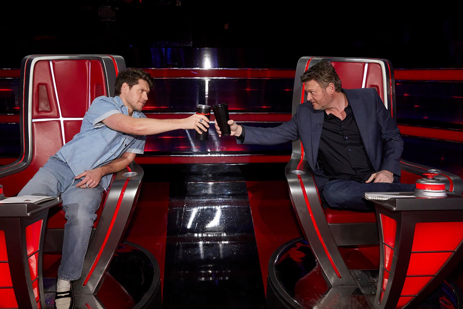Niall Horan and Blake Shelton toast with their cups as they sit in their chairs on The Voice Season 23
