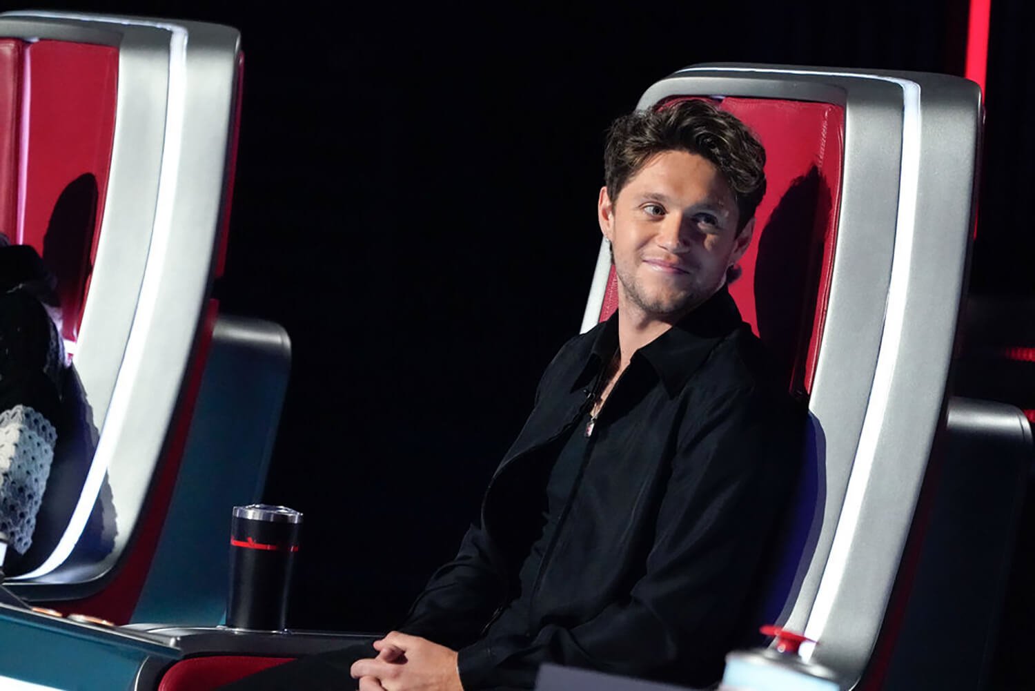 Niall Horan smiles in his seat and looks to the side on The Voice Season 23