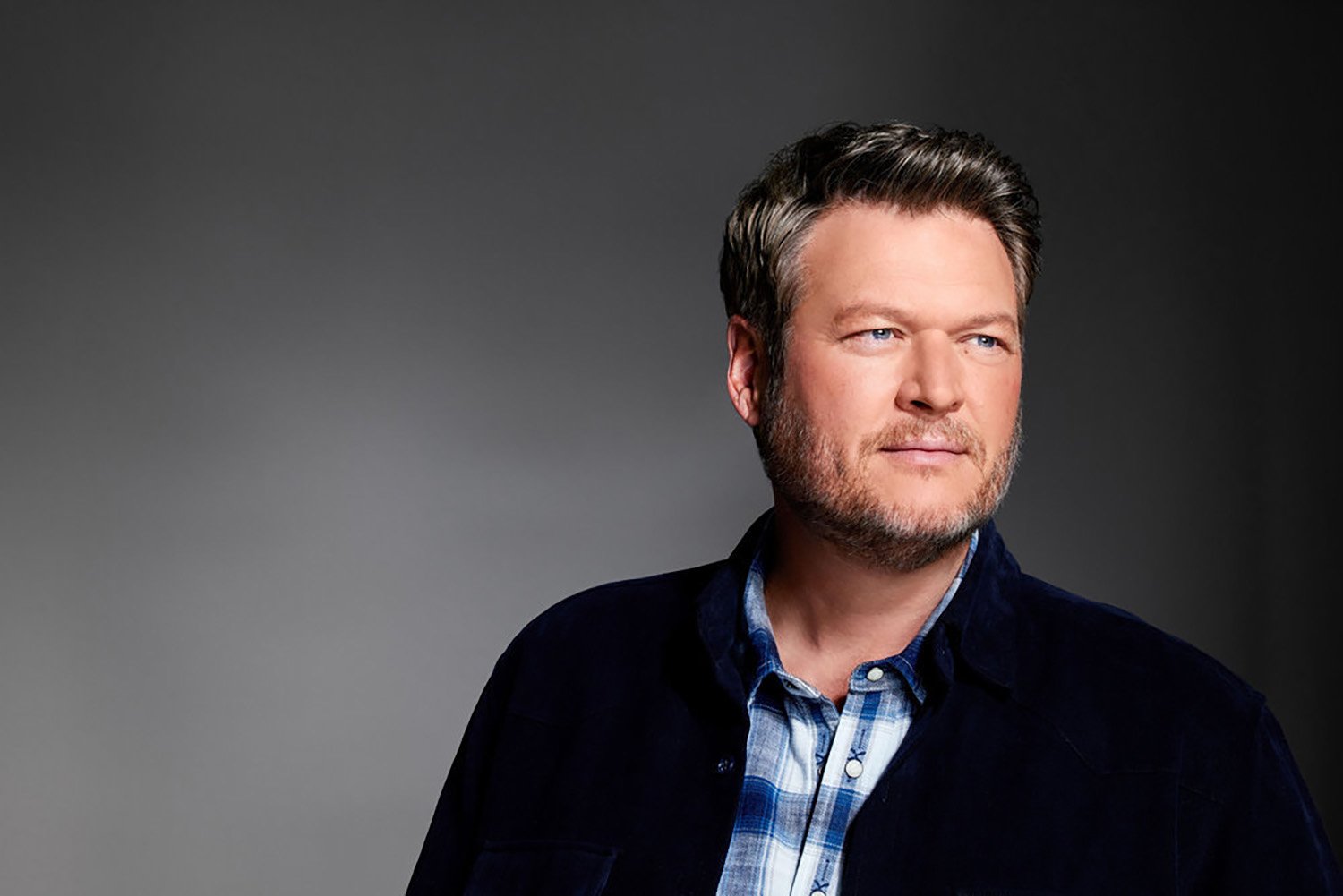 Blake Shelton, wearing a blue plaid flannel and a black jacket, poses against a gray backdrop for The Voice Season 23.