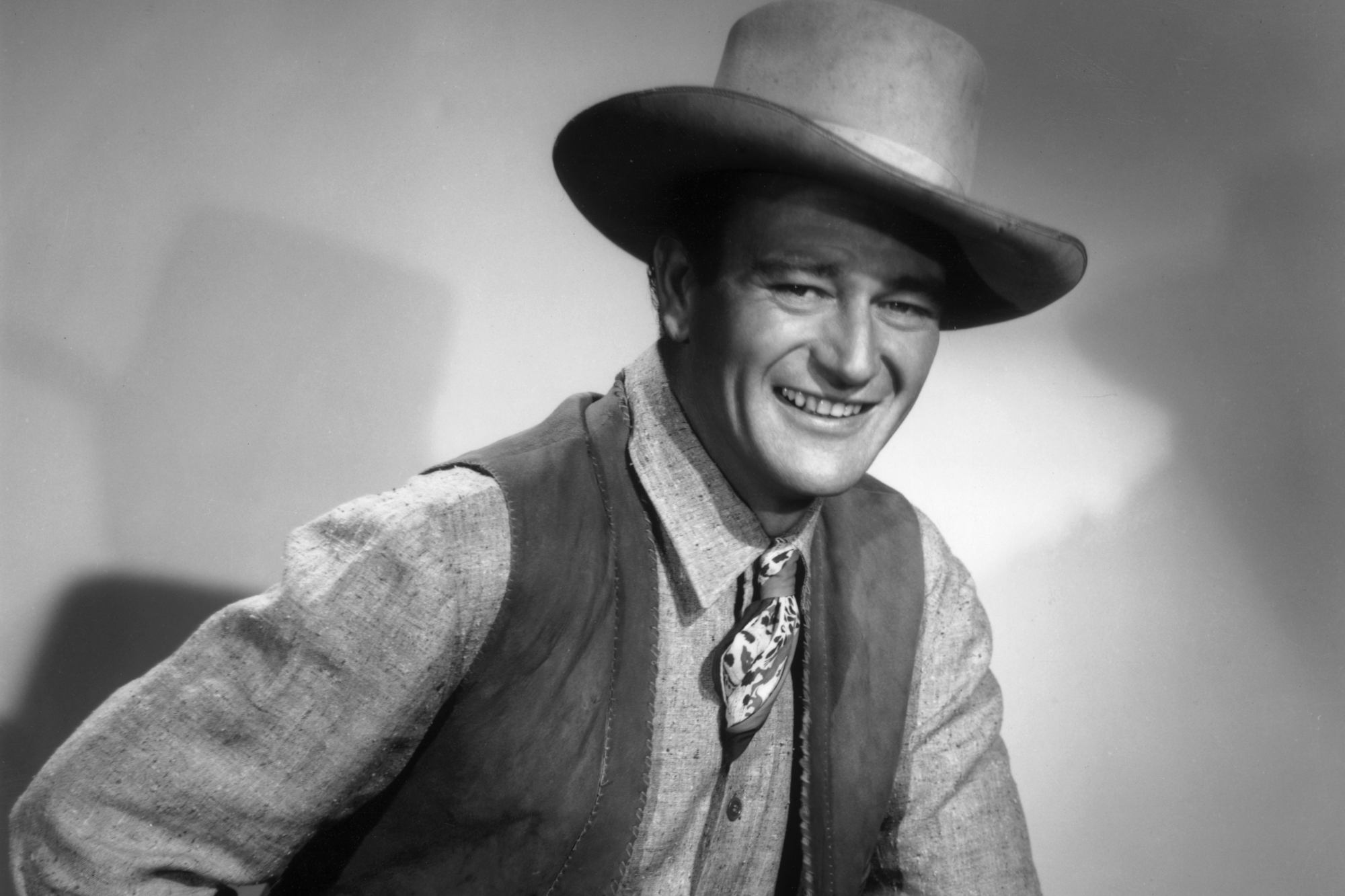 Actor and once stuntman John Wayne smiling in a black-and-white portrait, wearing Western clothing.
