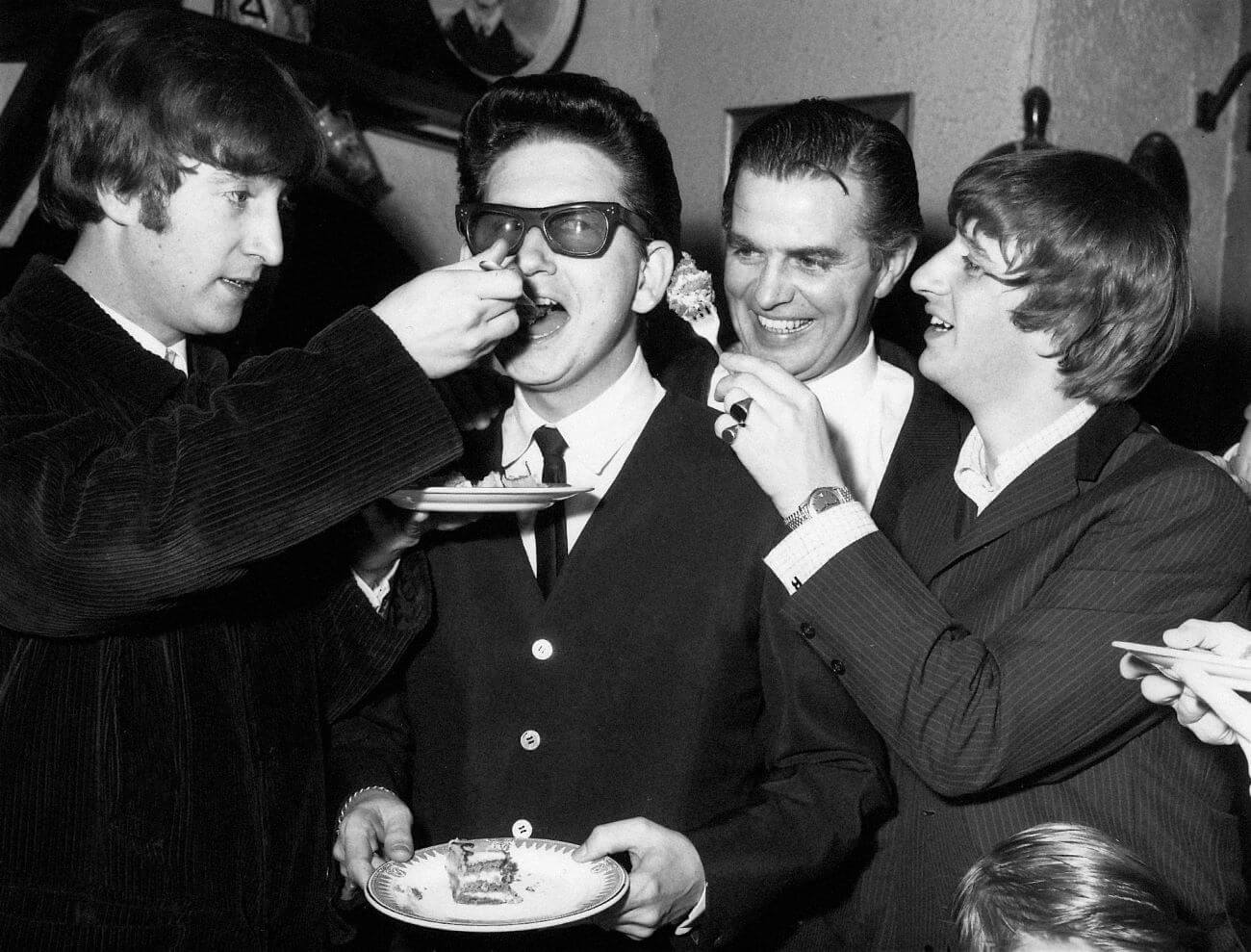 A black and white picture of Beatles John Lennon and Ringo Starr feeding Roy Orbison cake.