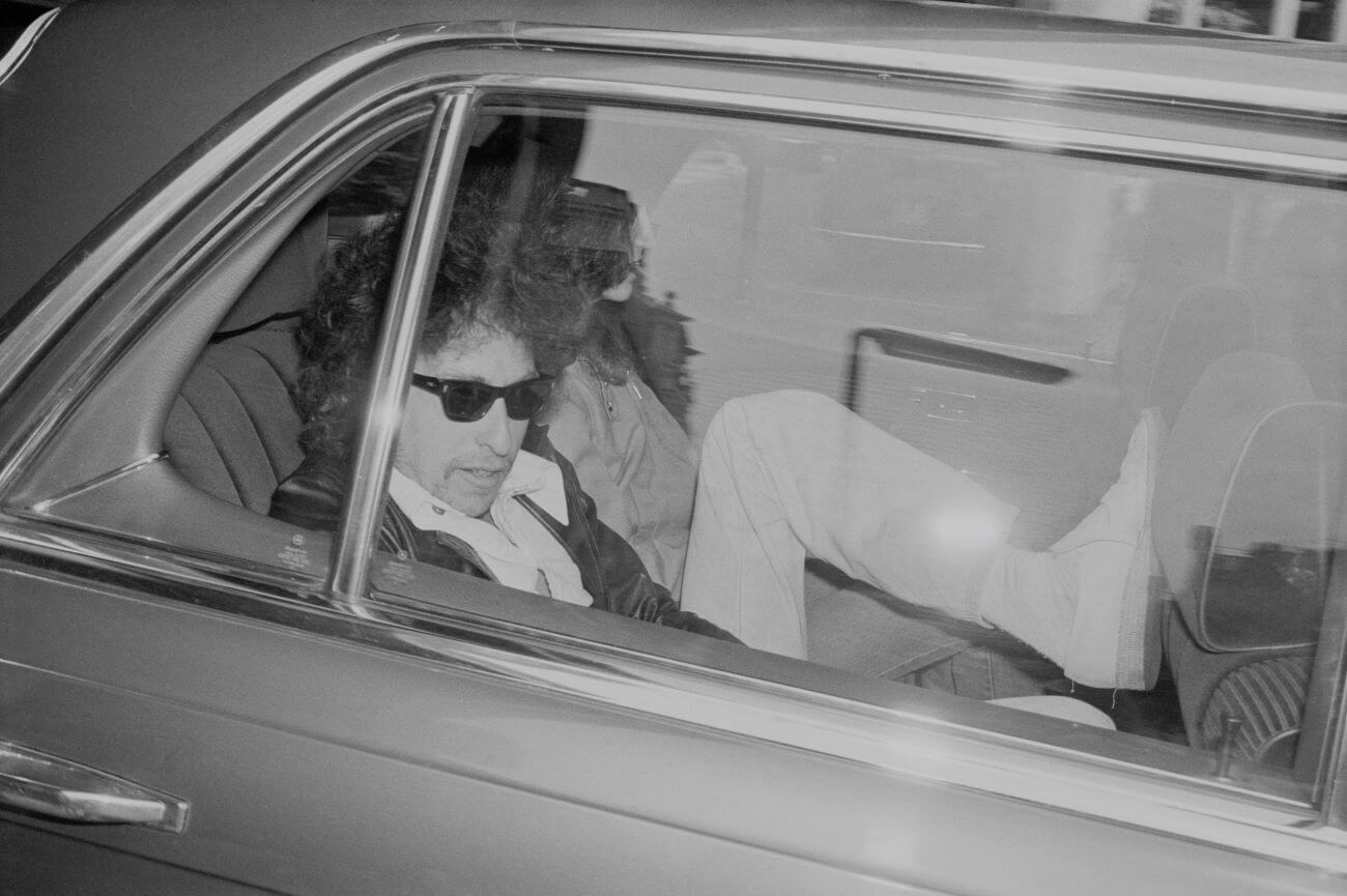 A black and white picture of Bob Dylan wearing sunglasses in the back seat of a car. His foot rests against the seat in front of him.