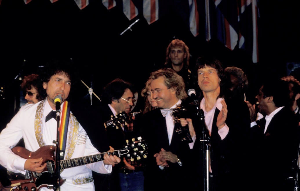 Bob Dylan plays a guitar and Mick Jagger claps his hands. They both stand in front of microphones. 