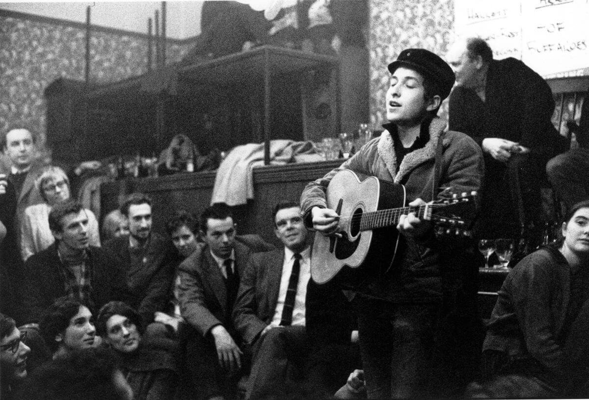 Bob Dylan plays guitar in a room full of people in the Sixties. 